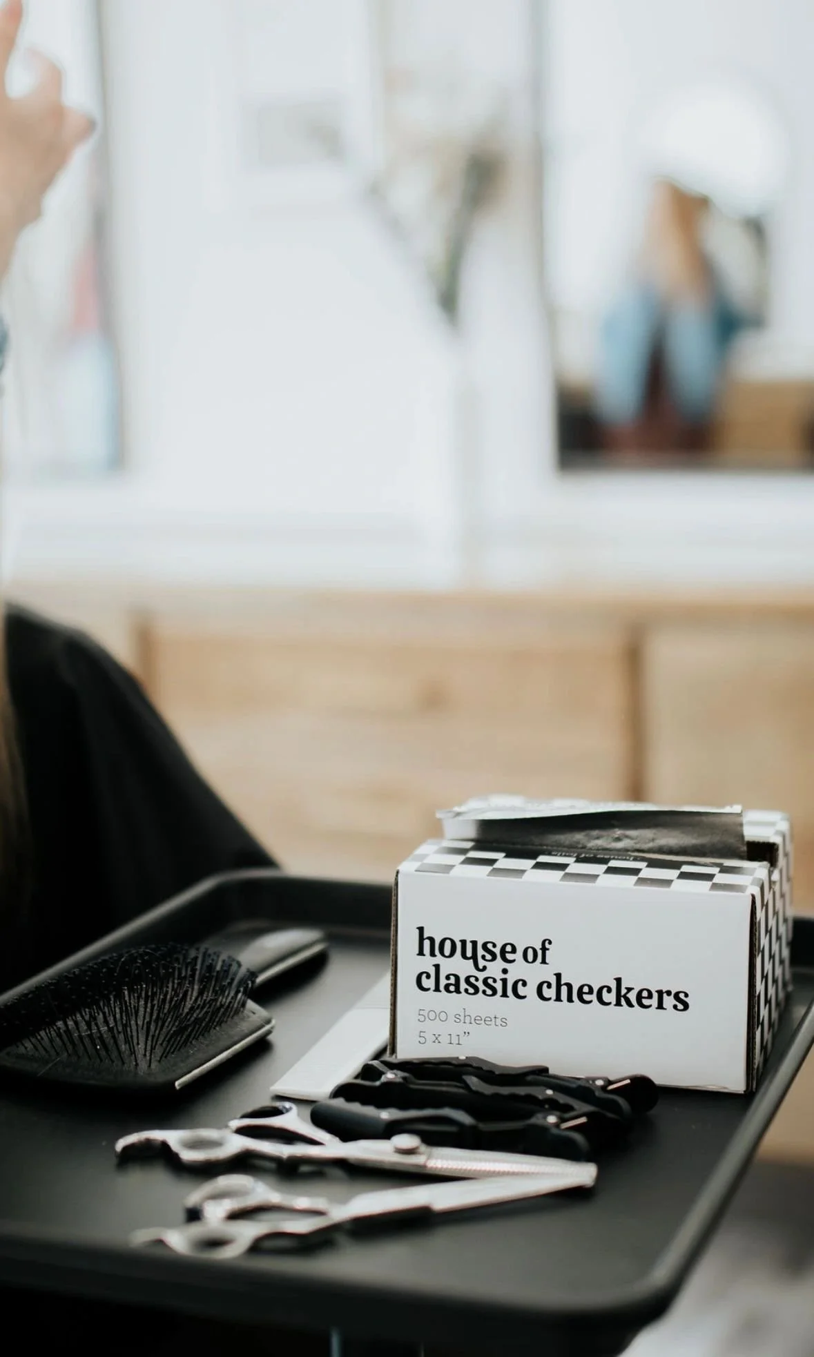 A black tray holding hairdressing tools including a hairbrush, a pair of scissors, and a smaller tool, with a box labeled 'house of classic checkers' containing 500 sheets of checkered paper in the background.
