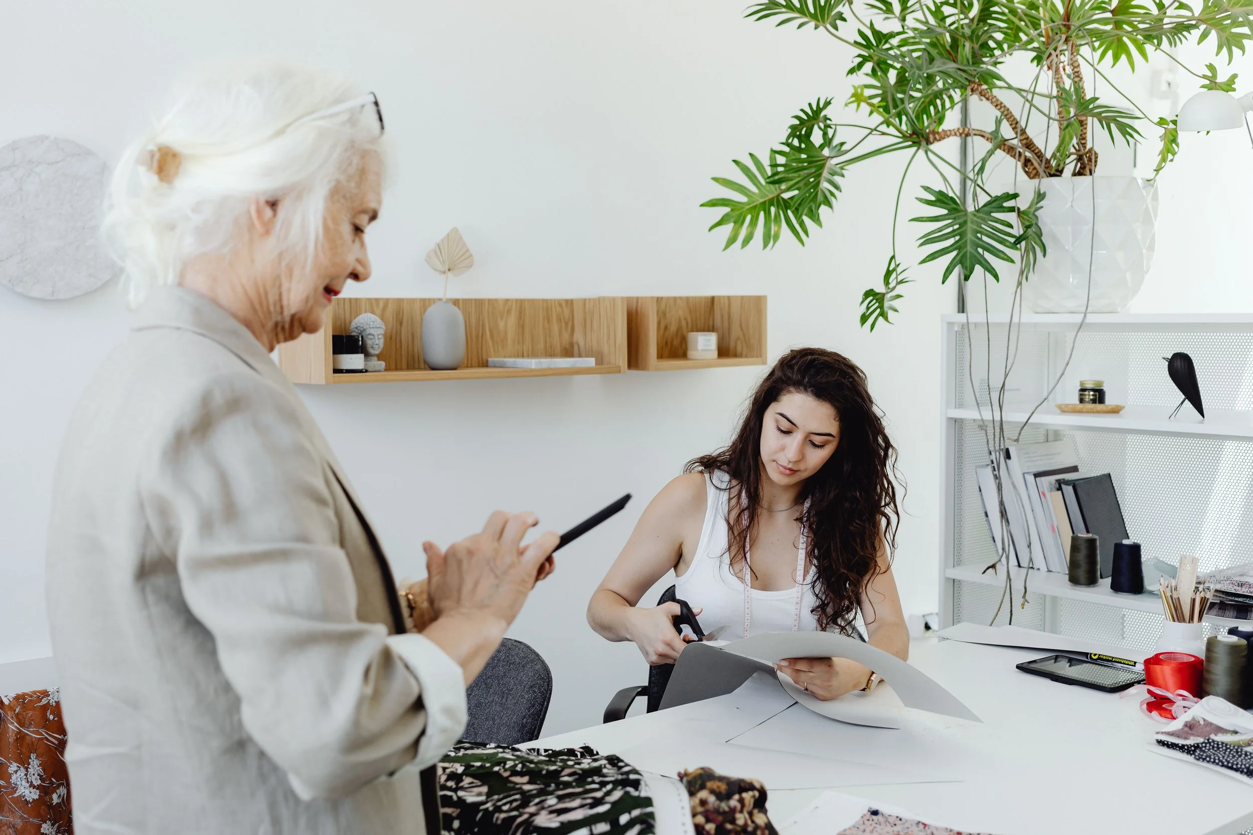 An elderly woman and a young woman in a bright, modern workspace involving sewing or craft work, with various materials and plants visible.