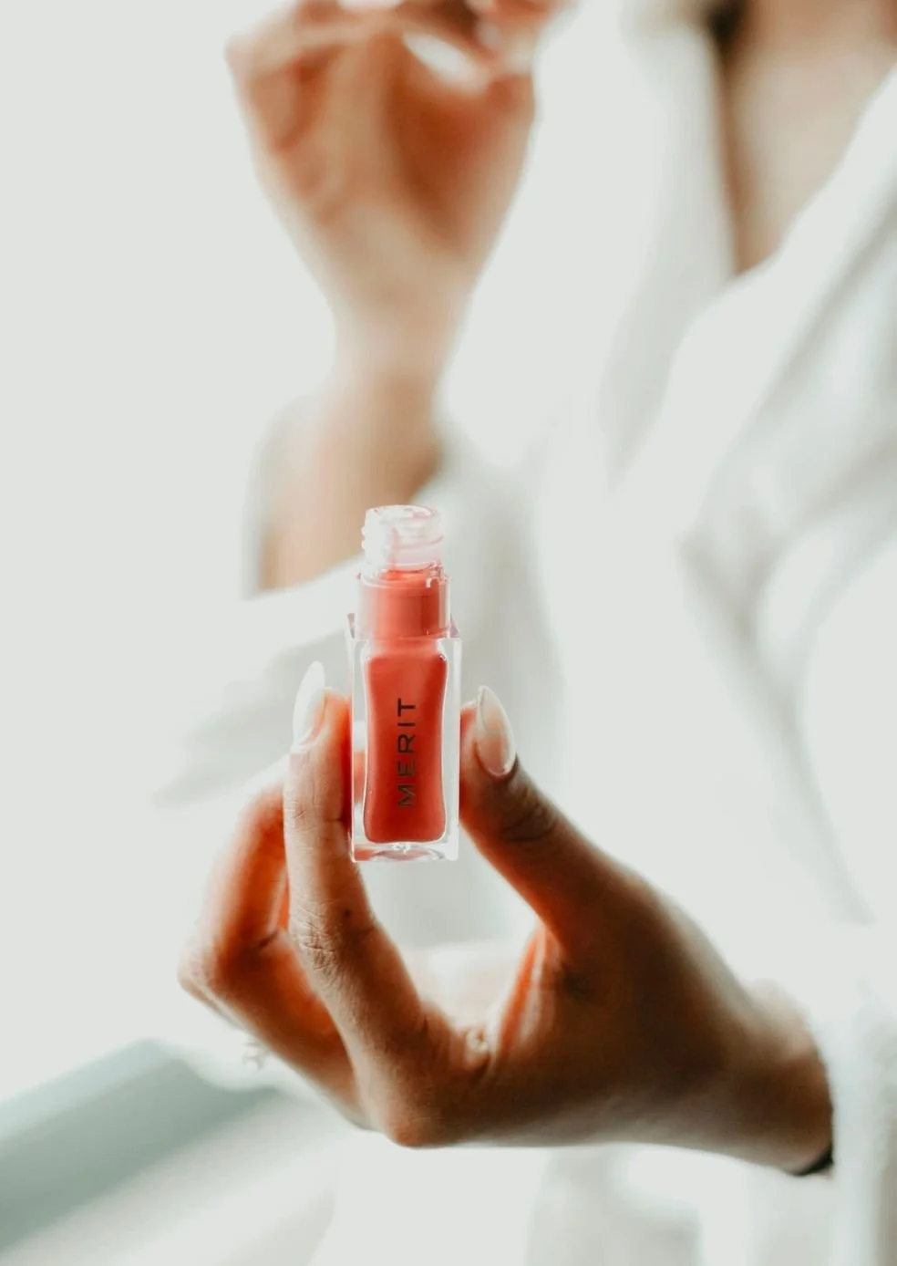 Close-up of a hand holding a coral-colored lip gloss tube labeled 'MERIT', with a blurred person in white in the background.