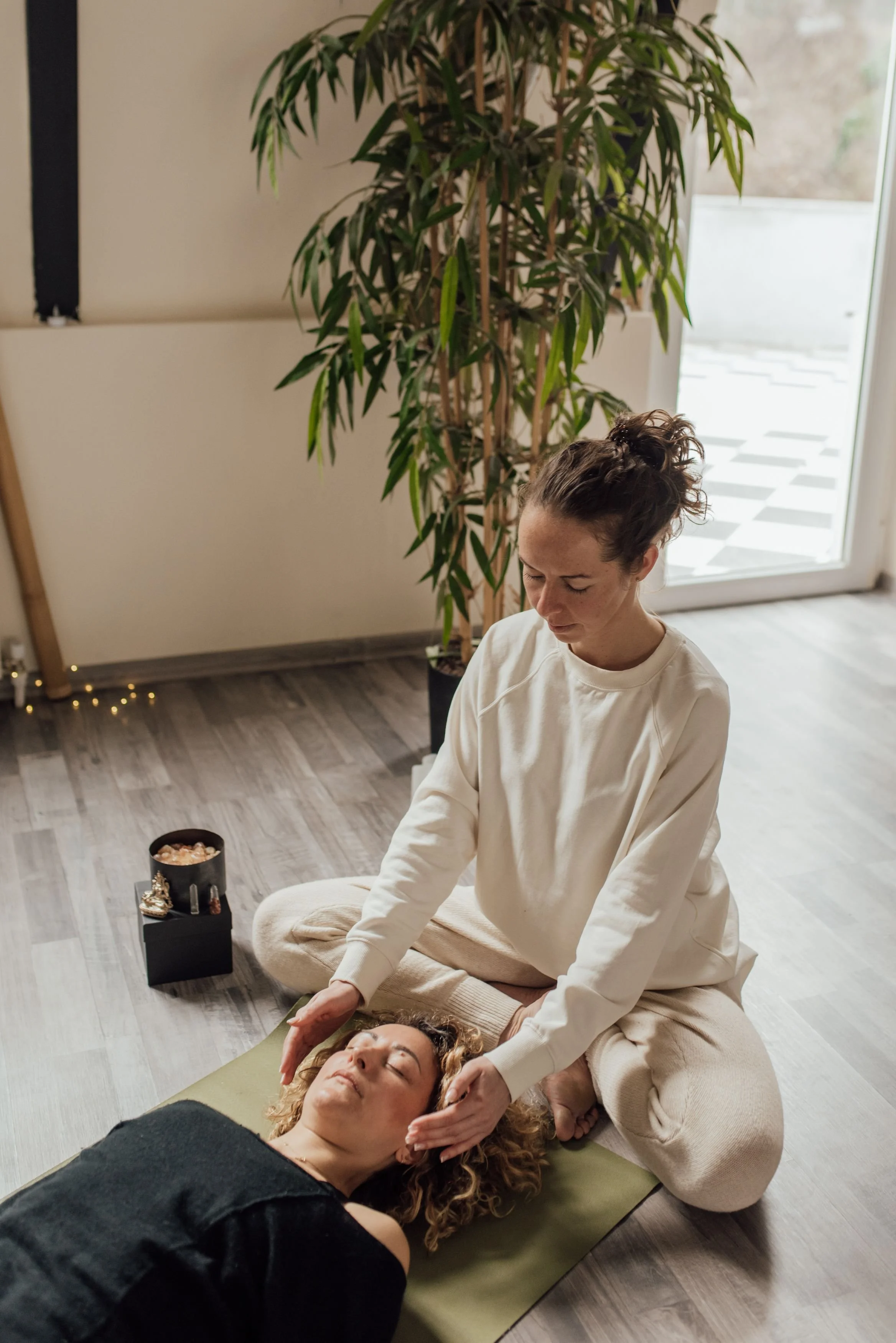 A woman receiving a facial massage while lying on a yoga mat indoors. She has curly hair and dark clothing. Another woman, sitting cross-legged behind her, is providing the massage, dressed in a white long-sleeve shirt and beige pants. A small black box with jewelry sits nearby, and large green plants are in the background near a glass door.