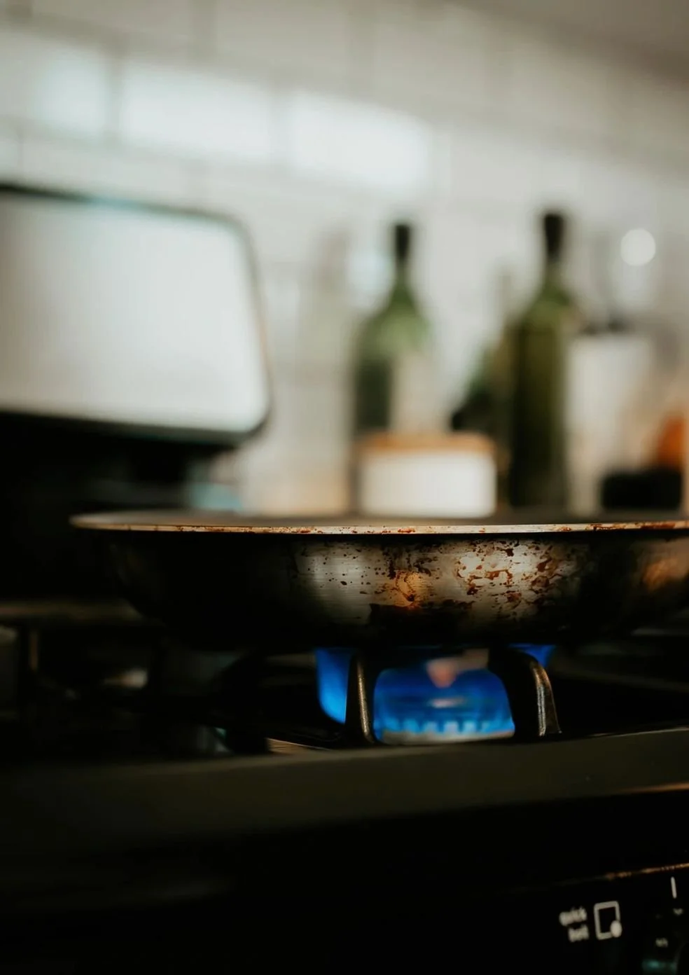 Close-up of a lit gas stove burner with a blue flame, with a worn frying pan on top, in a kitchen setting.