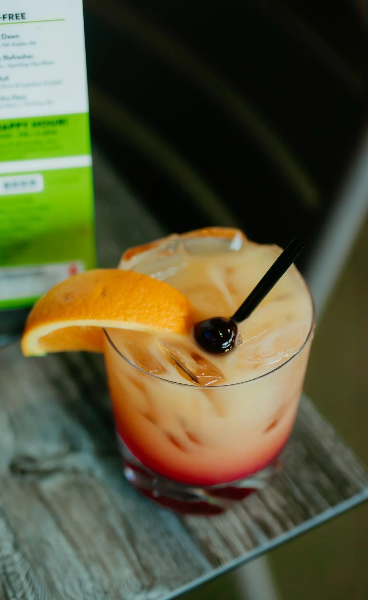 A tropical cocktail with an orange slice and a cherry, served over ice with a black straw on a wooden surface.