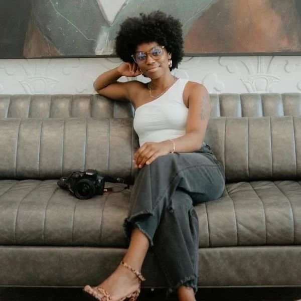 A woman with curly black hair, glasses, and earrings sitting on a gray couch with a camera beside her. She is wearing a white sleeveless top, dark jeans, and heels, posing with her left hand resting on her head in a relaxed manner.