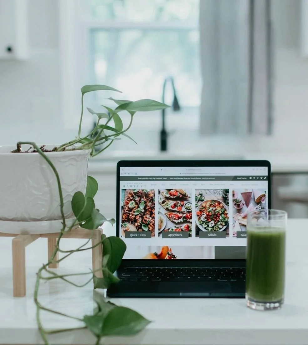 Laptop displaying a website with food images, a potted plant on a small stand, and a green smoothie on a white table in a bright room with a window in the background.