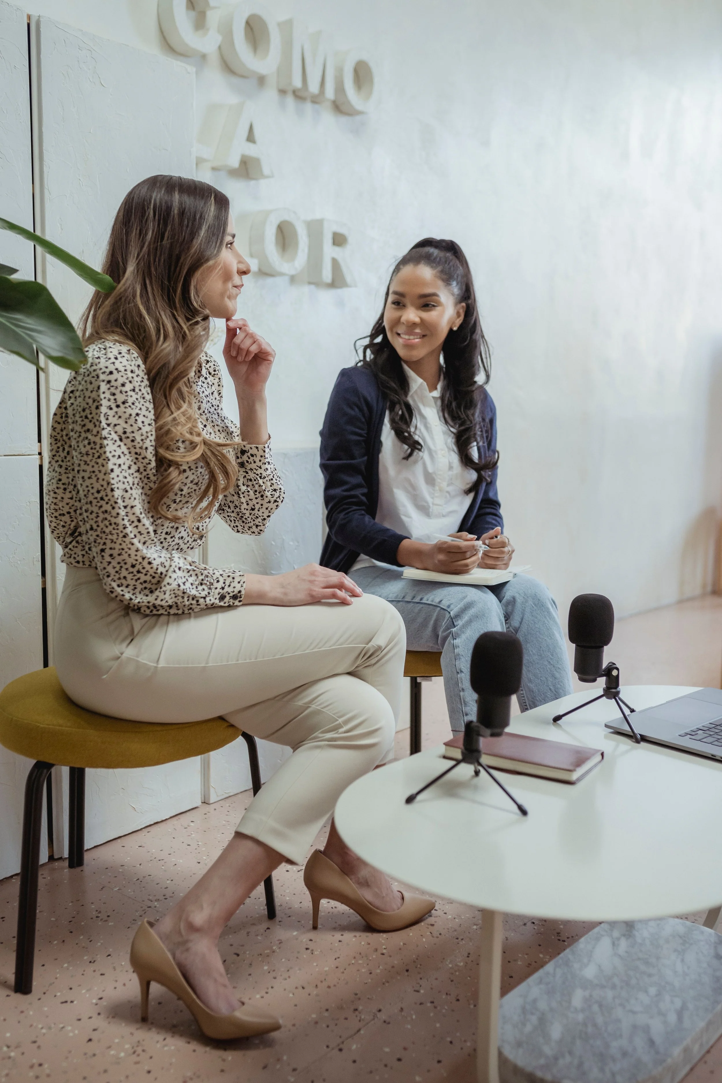 Two women engaged in a conversation during a podcast recording session, sitting at a round table with microphones, notebooks, and a laptop, in a room with white walls and decorative letters spelling 'Come As For' in the background.