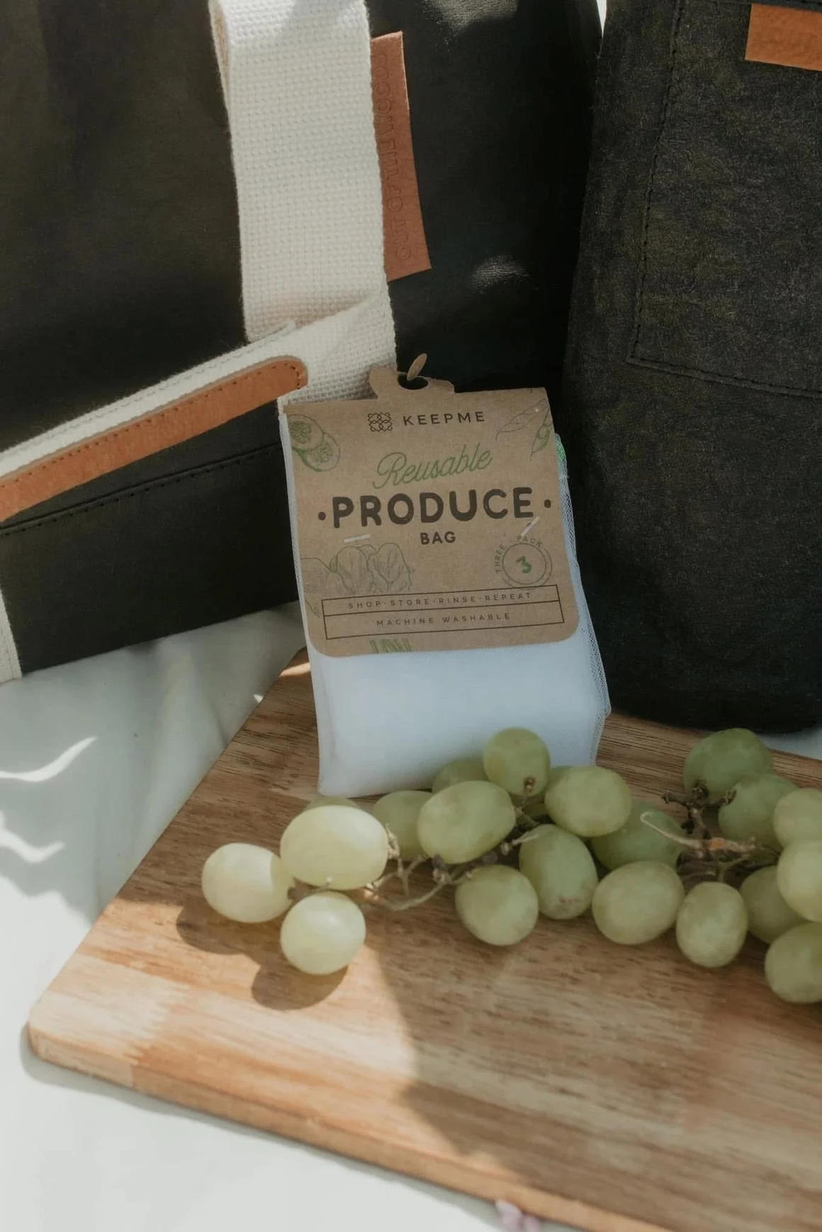 A reusable produce bag filled with produce on a wooden cutting board, with a bunch of green grapes nearby, and bags in the background.