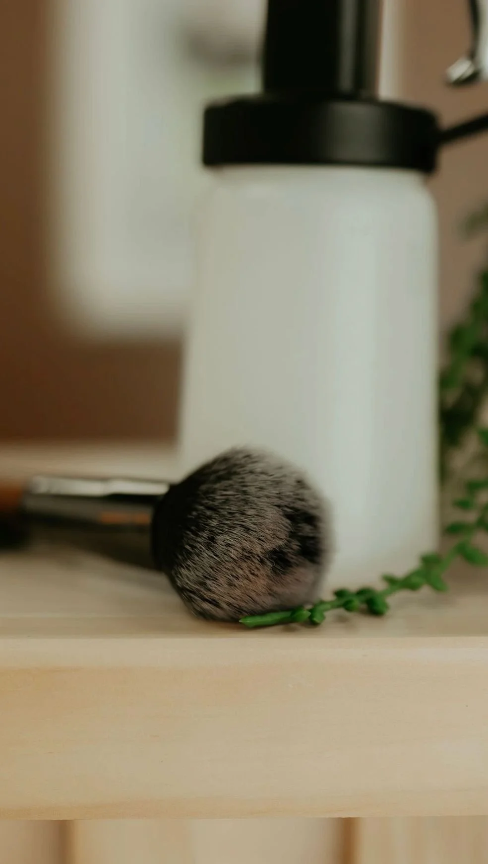 A fluffy makeup brush with black and gray bristles resting on a wooden surface next to an artificial green plant, with a blurred white container and other objects in the background.