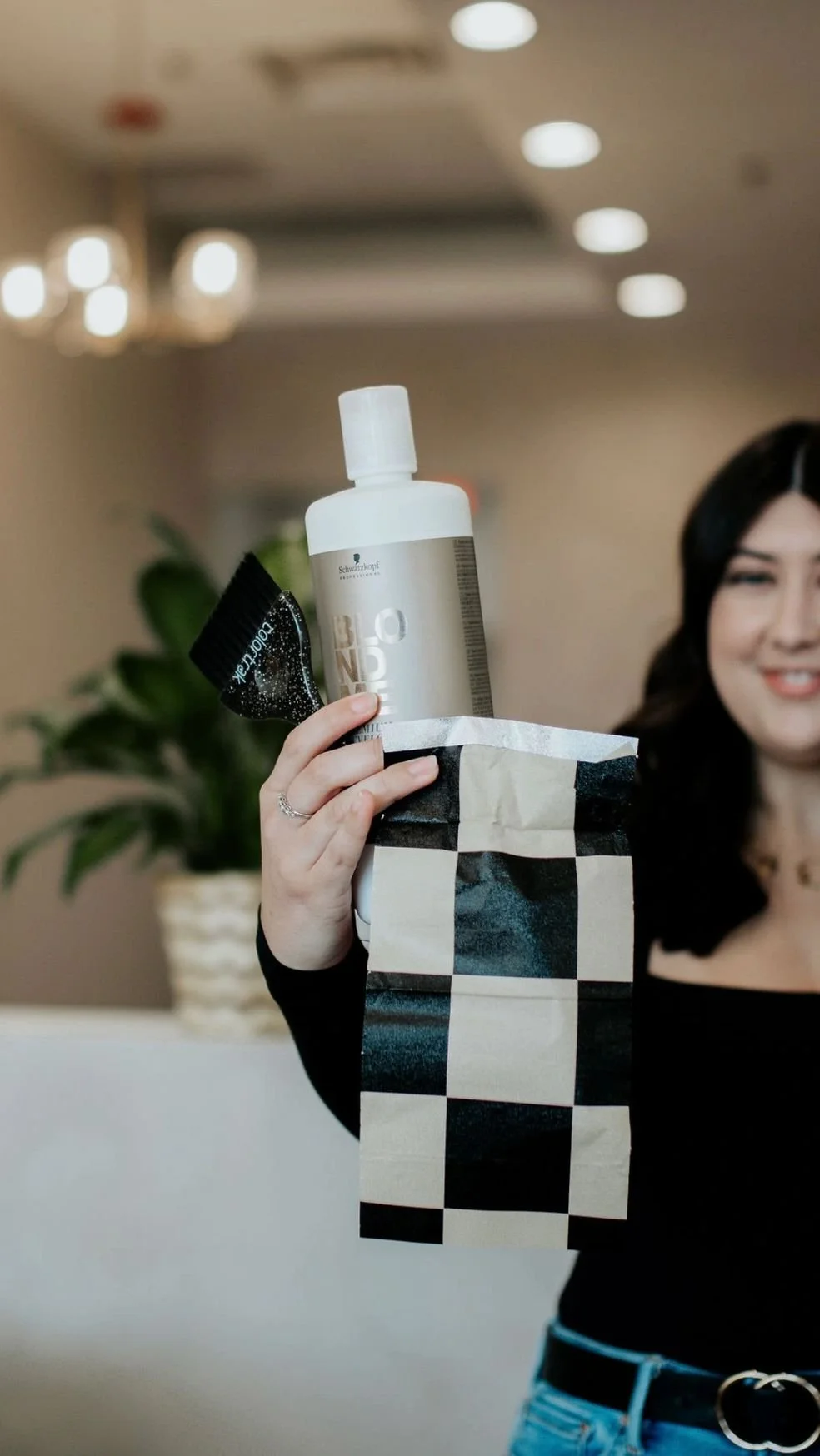 A woman holding a checkered paper bag with hair and some hair care products visible from the top, including a bottle, a tube, and a container.