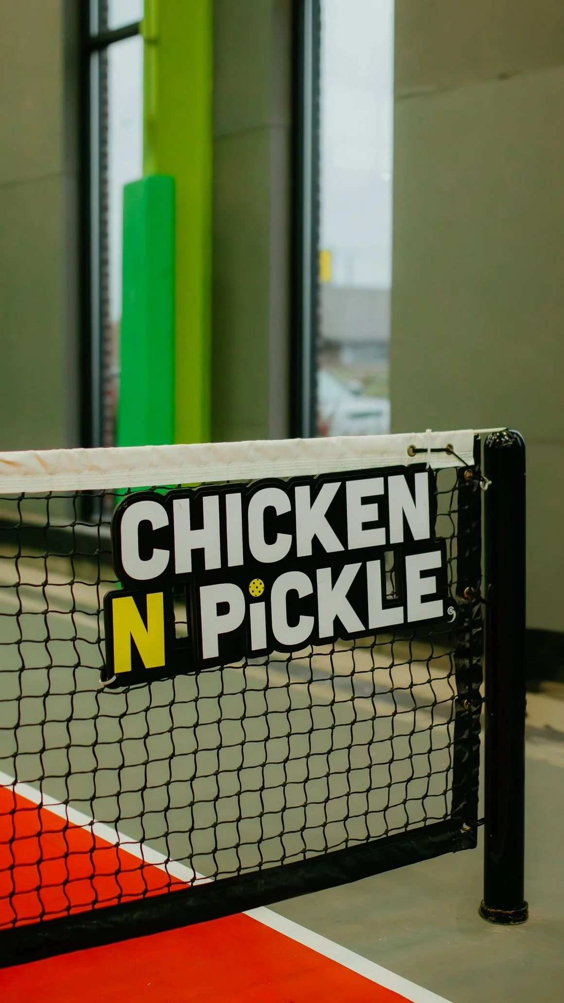 Sign on a fence that reads 'Chicken N Pickle' with the letters N in yellow and the rest in black and white, on a pickleball court.