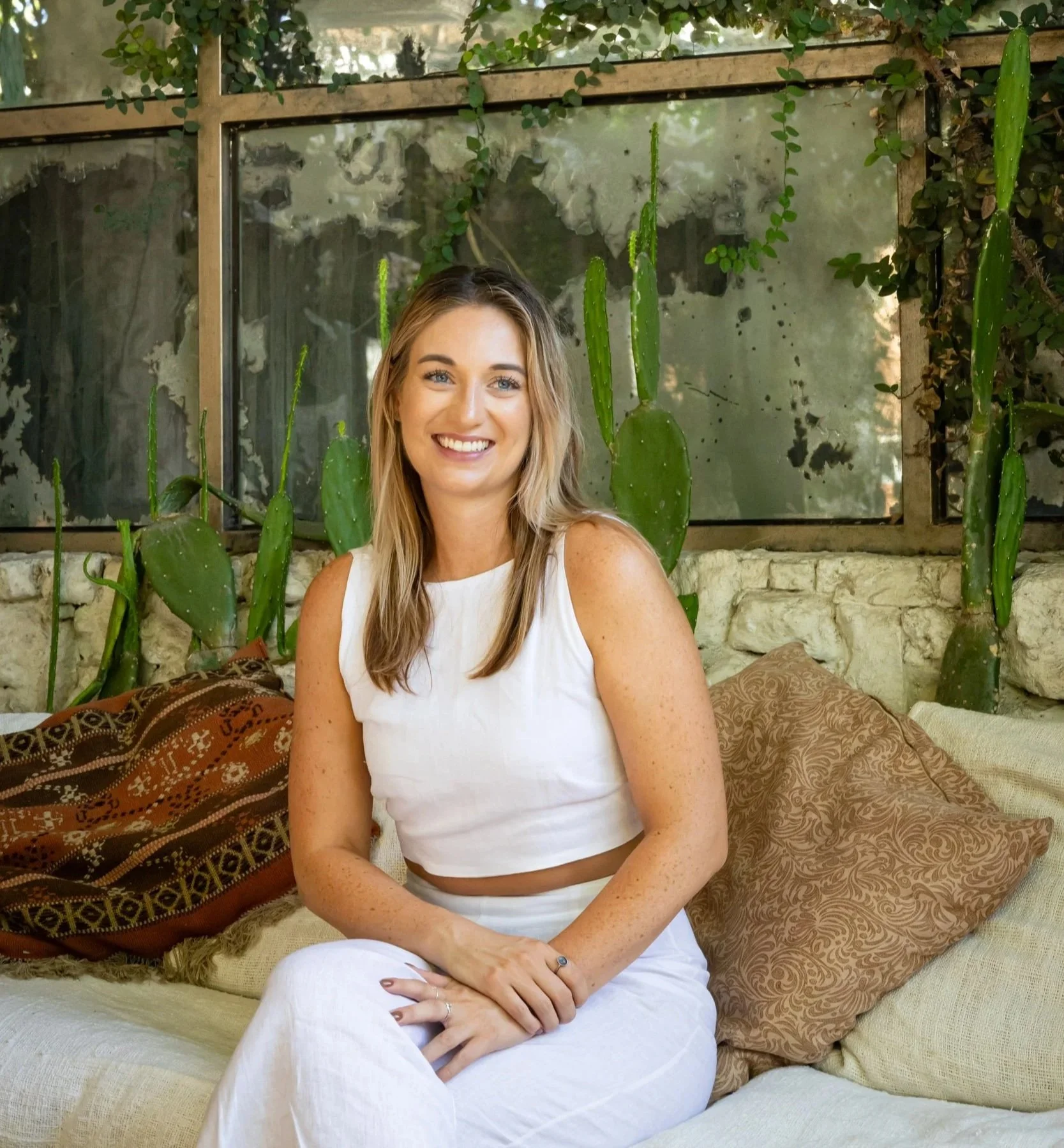 A smiling woman with blonde hair and blue eyes wearing a sleeveless white top and white pants, sitting on a couch with decorative pillows in a room with tall cactus plants and a rustic window in the background.