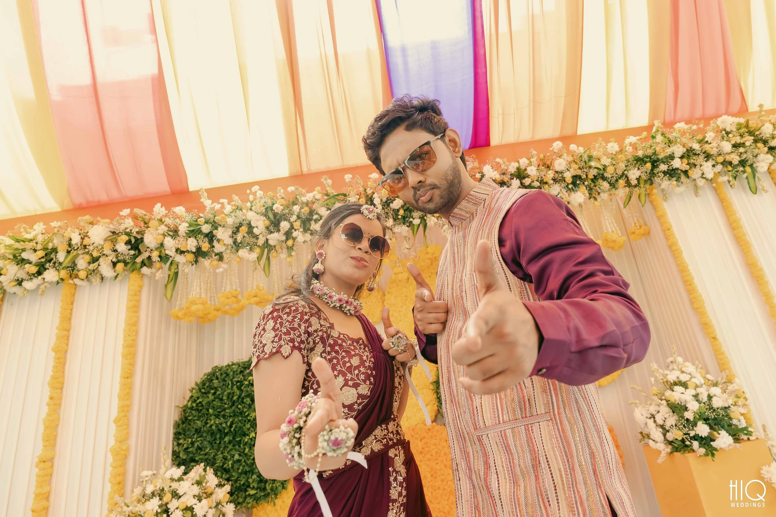 A couple dressed in traditional Indian wedding attire striking a pose at a decorated wedding stage with colorful drapes and floral arrangements.