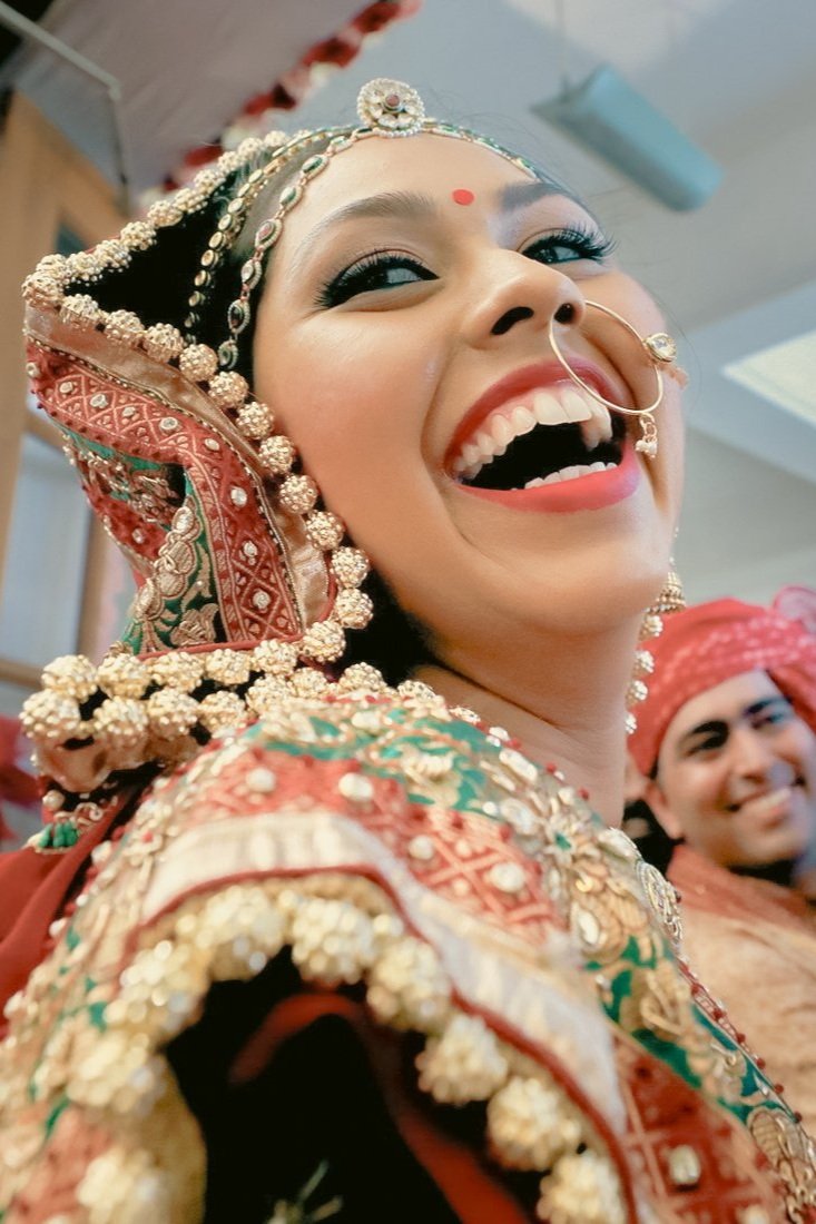A woman dressed in traditional Indian wedding attire, smiling widely and wearing elaborate jewelry and a decorated headpiece, with a man in similar attire smiling in the background.