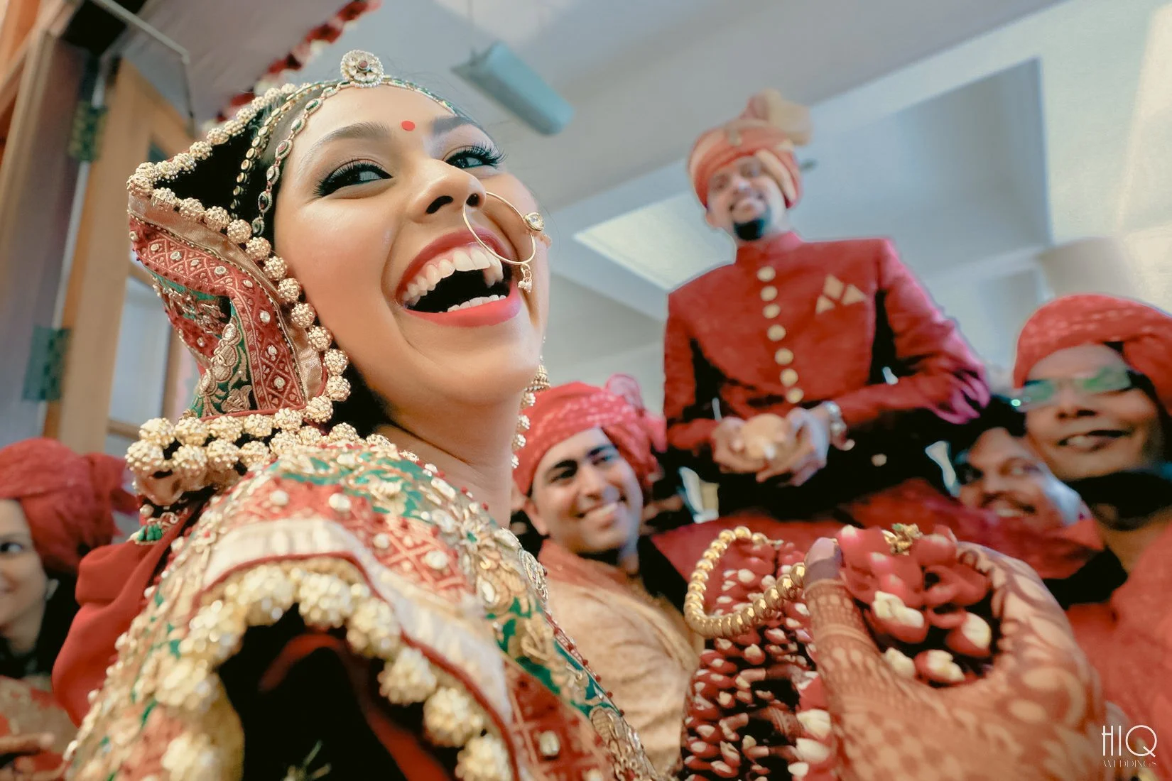 A bride dressed in traditional Indian wedding attire, smiling and surrounded by relatives wearing red turbans, in a festive celebration.
