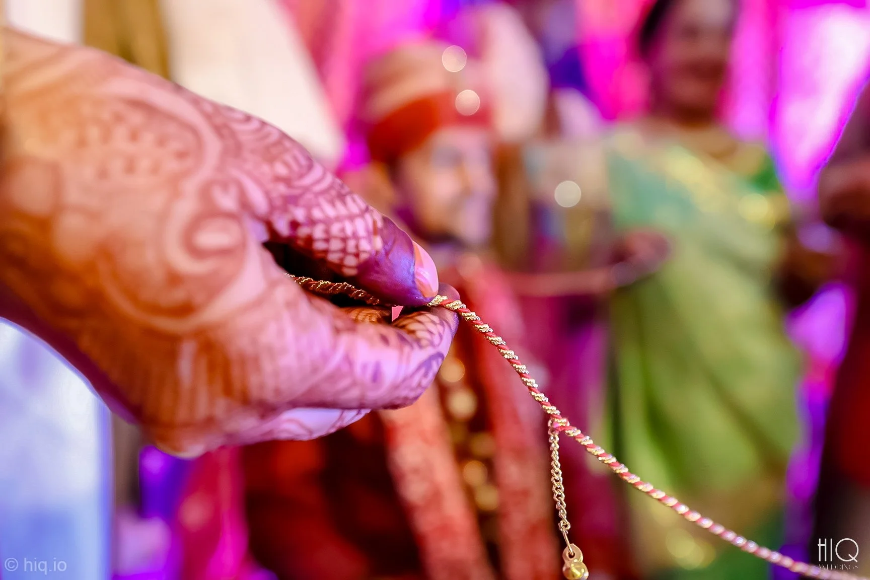 Close-up of a hand holding a gold necklace during a colorful Indian wedding ceremony, with women in traditional attire in the background.