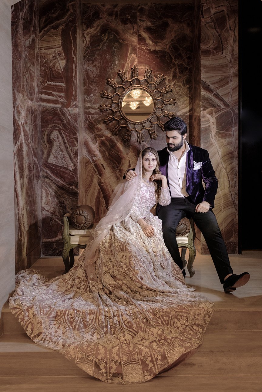 A bride and groom sitting on a vintage chair in a luxurious room with marble walls and a round mirror with a sunburst frame captured by HiQ Weddings