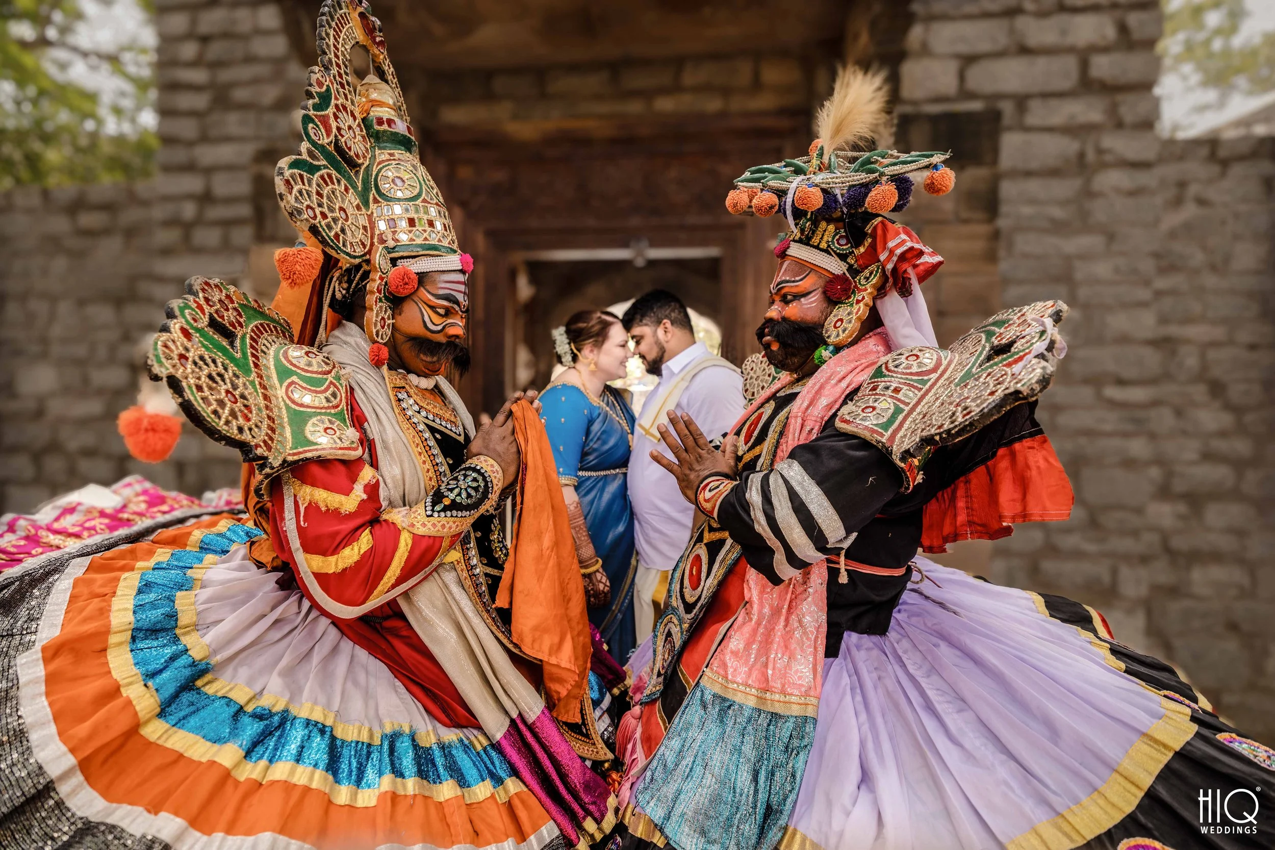 Two men dressed in traditional Indian costumes performing a dance facing each other, with a wedding couple in traditional attire in the background.