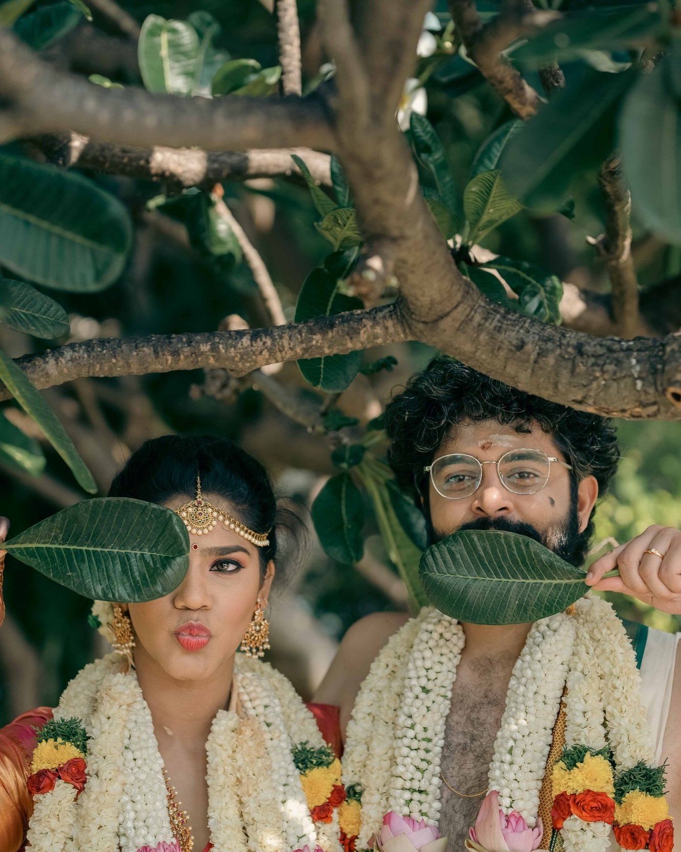 A couple in traditional Indian wedding attire, wearing floral garlands, posing beneath a tree. The woman is making a playful pout, and the man is holding a leaf in front of his face.