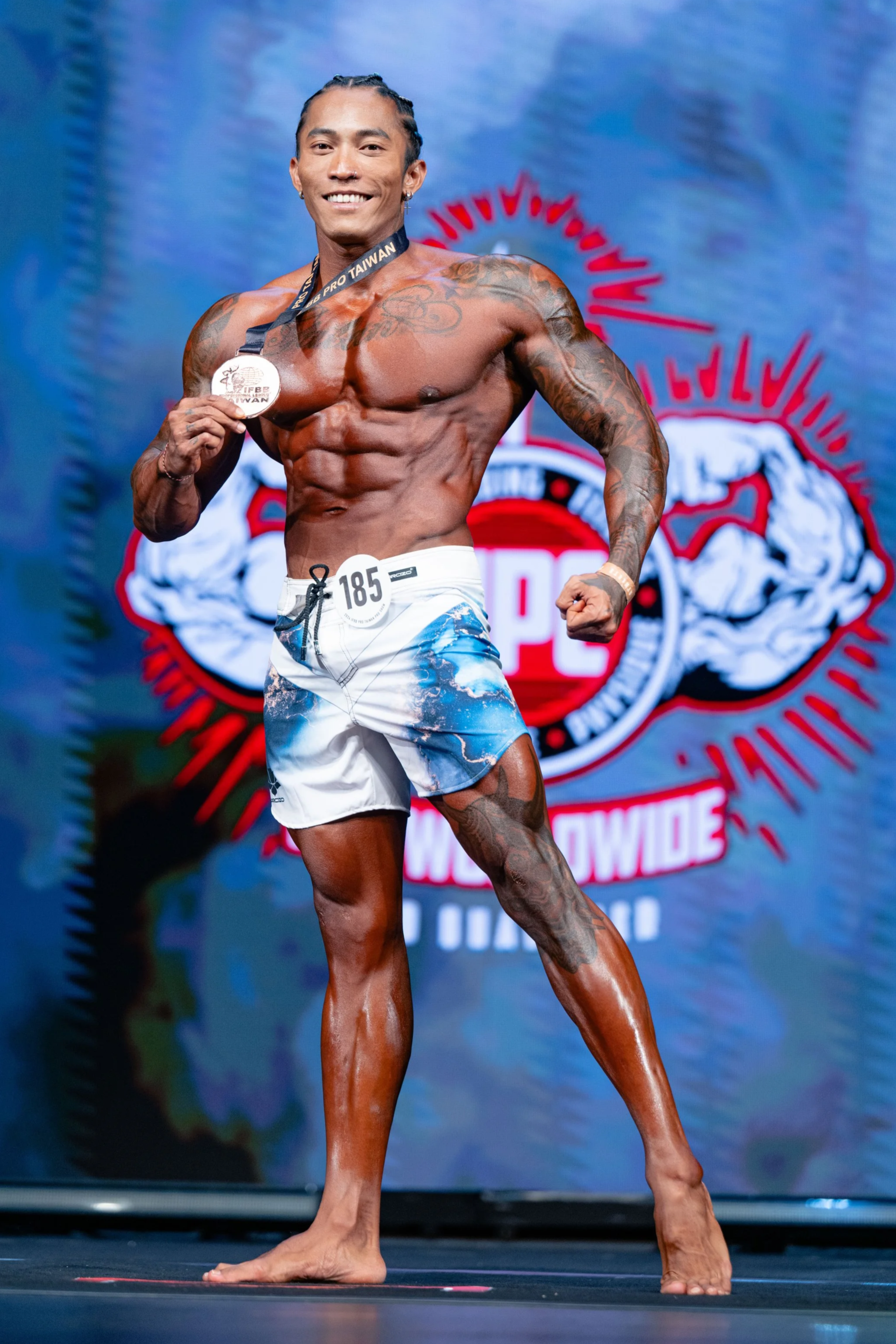 A muscular man with tattoos poses confidently on stage at a bodybuilding competition, holding a medal while showcasing his physique. Set against a backdrop featuring a logo with muscular arms and the words “Worldwide” and “Pro Taiwan,” the moment cap