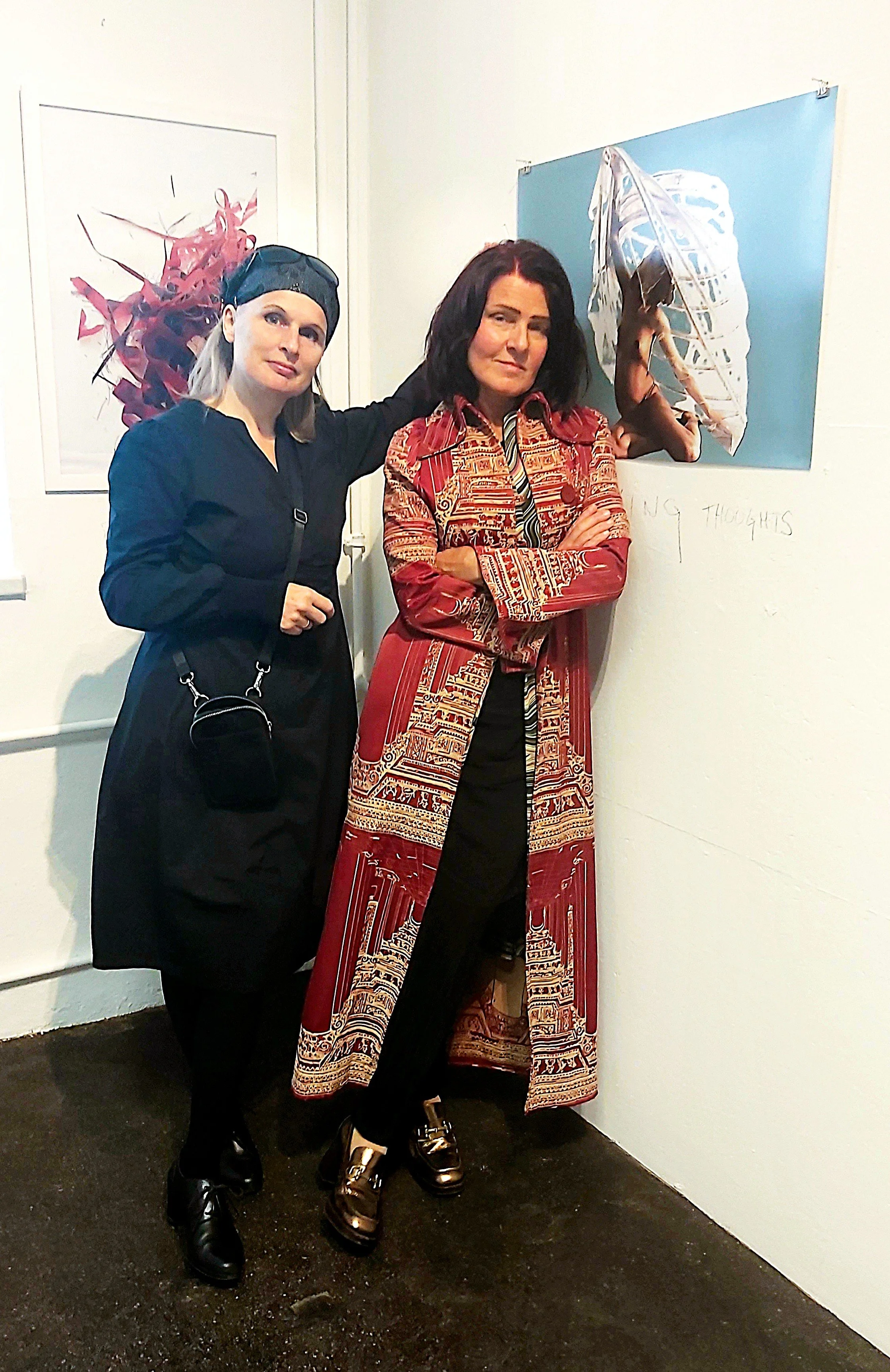 Zwei Frauen stehen nebeneinander in einer Galerie. Eine trägt eine schwarze Kleidung mit Kopfbedeckung, die andere trägt ein langes rotes Kleid mit Muster. Im Hintergrund sind Kunstwerke an der Wand zu sehen.