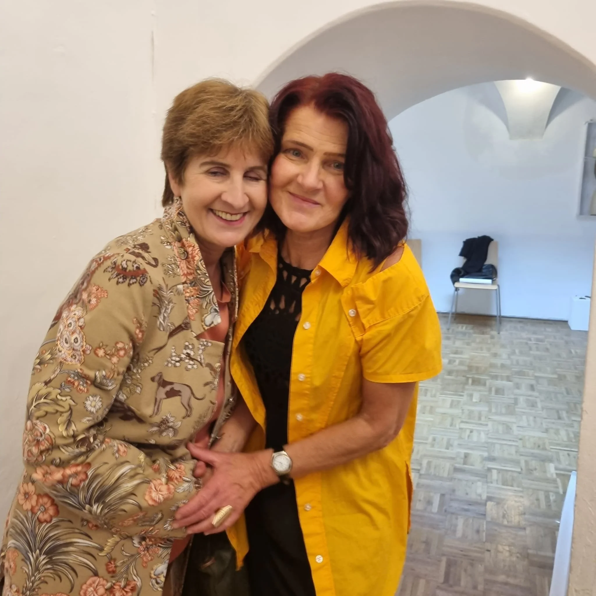 Zwei Frauen umarmen sich und lachen in einem Innenraum mit weißen Wänden und Steingartenboden.