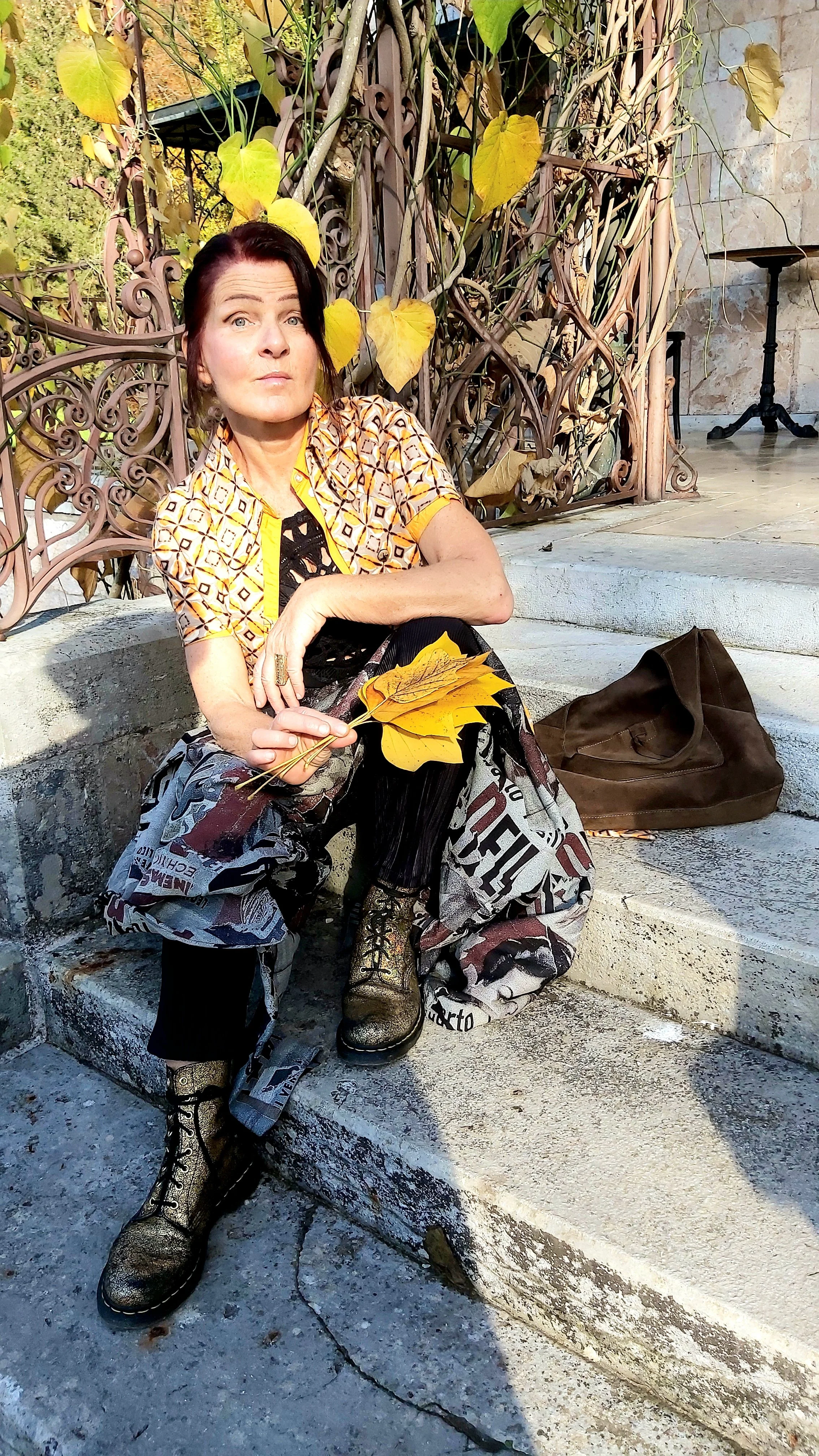 Marjeta Angerer-Guggenberger sitzt auf Stufen im Freien, hält gelbe Blätter in der Hand, trägt bunte Kleidung und schwarze Stiefel, neben einer braunen Tasche, im Hintergrund eine metallene Gartenfigur und eine Wand