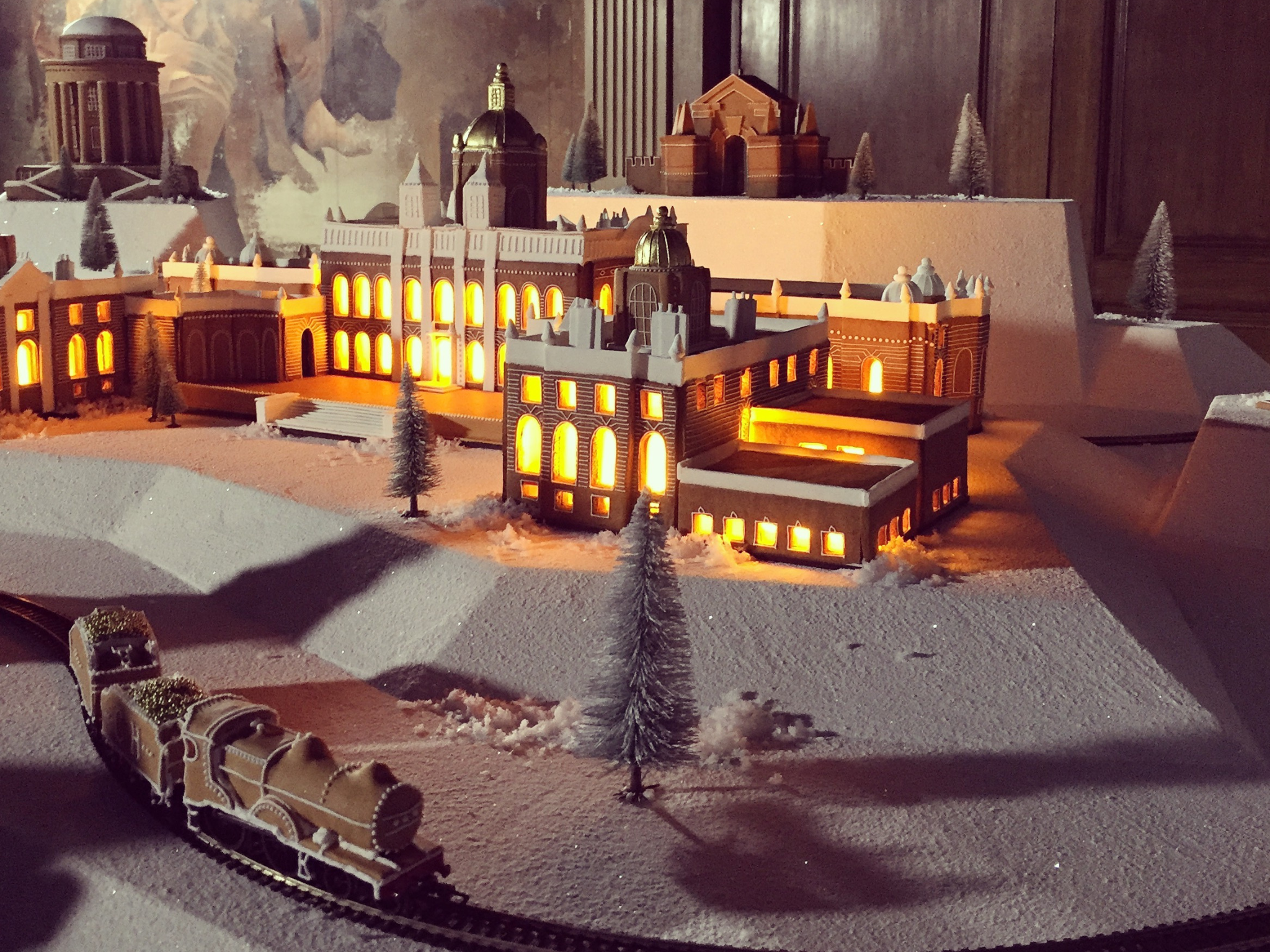A detailed miniature model of a large, illuminated gingerbread replica of Castle Howard with glowing windows, surrounded by snow-covered terrain, small pine trees, and a working gingerbread train in the foreground, set indoors.