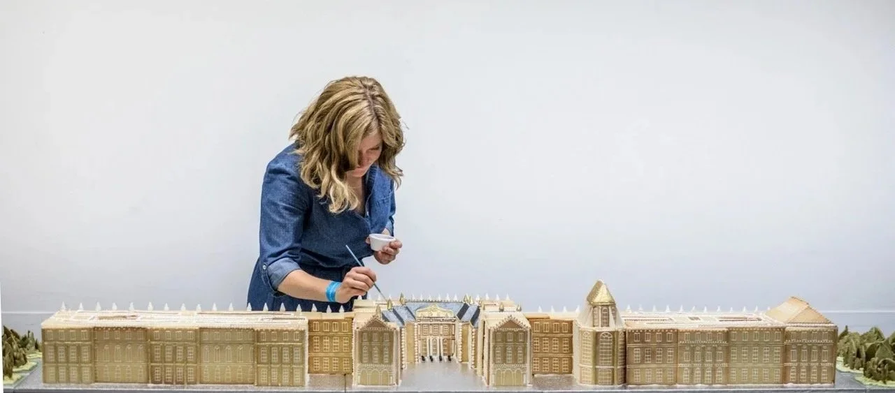 A woman with blonde hair wearing a blue shirt dress decorating a large gingerbread house resembling the Palace of Versailles.