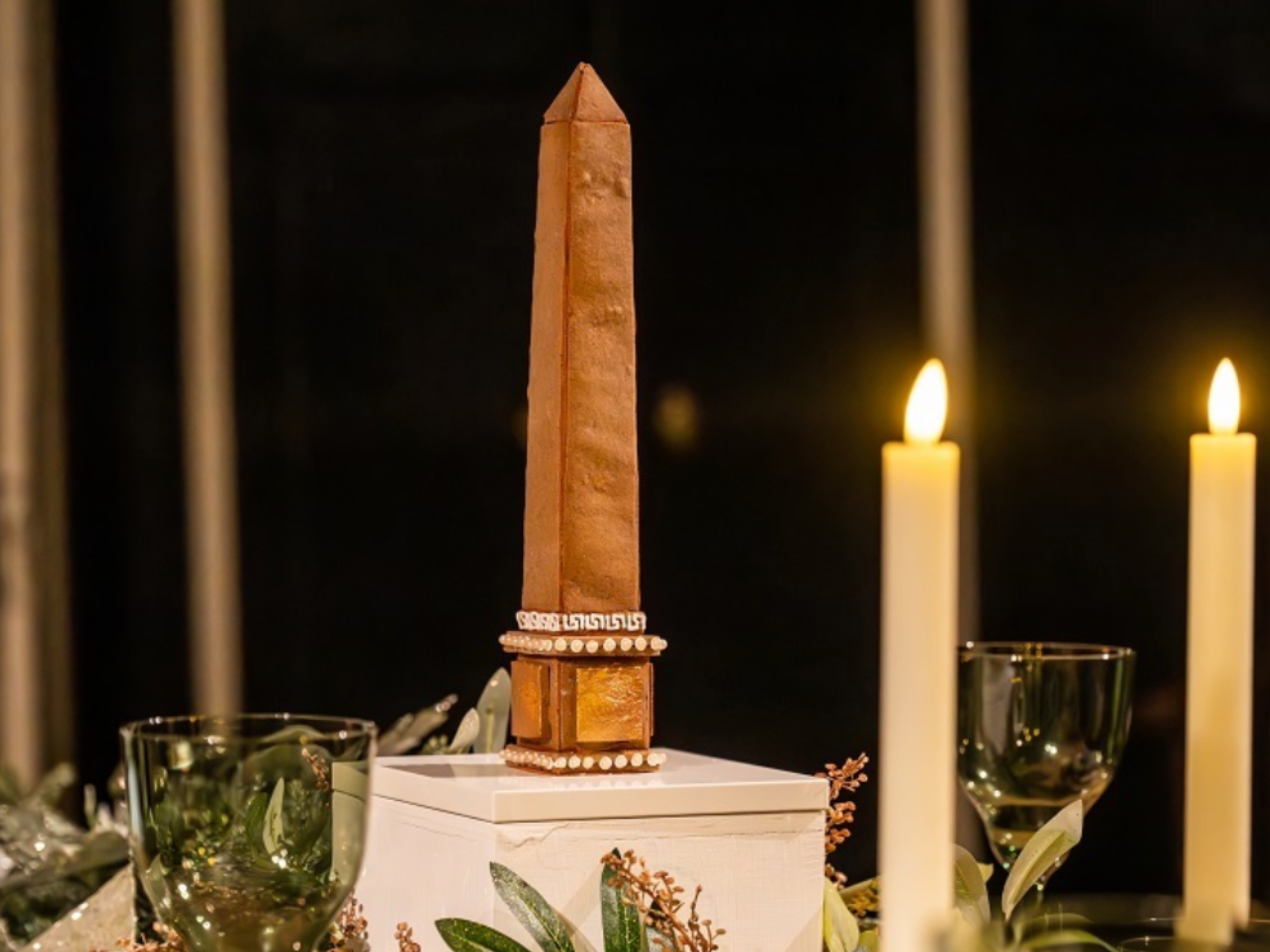 A tall, slim gingerbread obelisk with a pointed top, placed on a white pedestal, surrounded by leafy greenery, with two lit candles in the background. Part of a gingerbread installation of Holkham Hall.