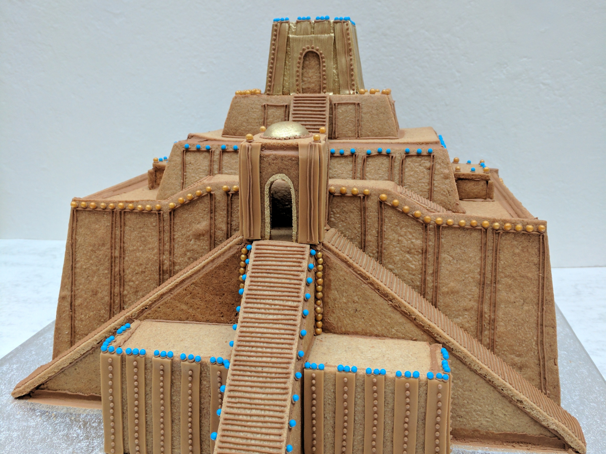A multi-tiered gingerbread monument decorated to resemble the Ziggurat of Ur, a stepped pyramid with stairs and architectural details, primarily in shades of brown and beige, with small blue and gold ball decorations.