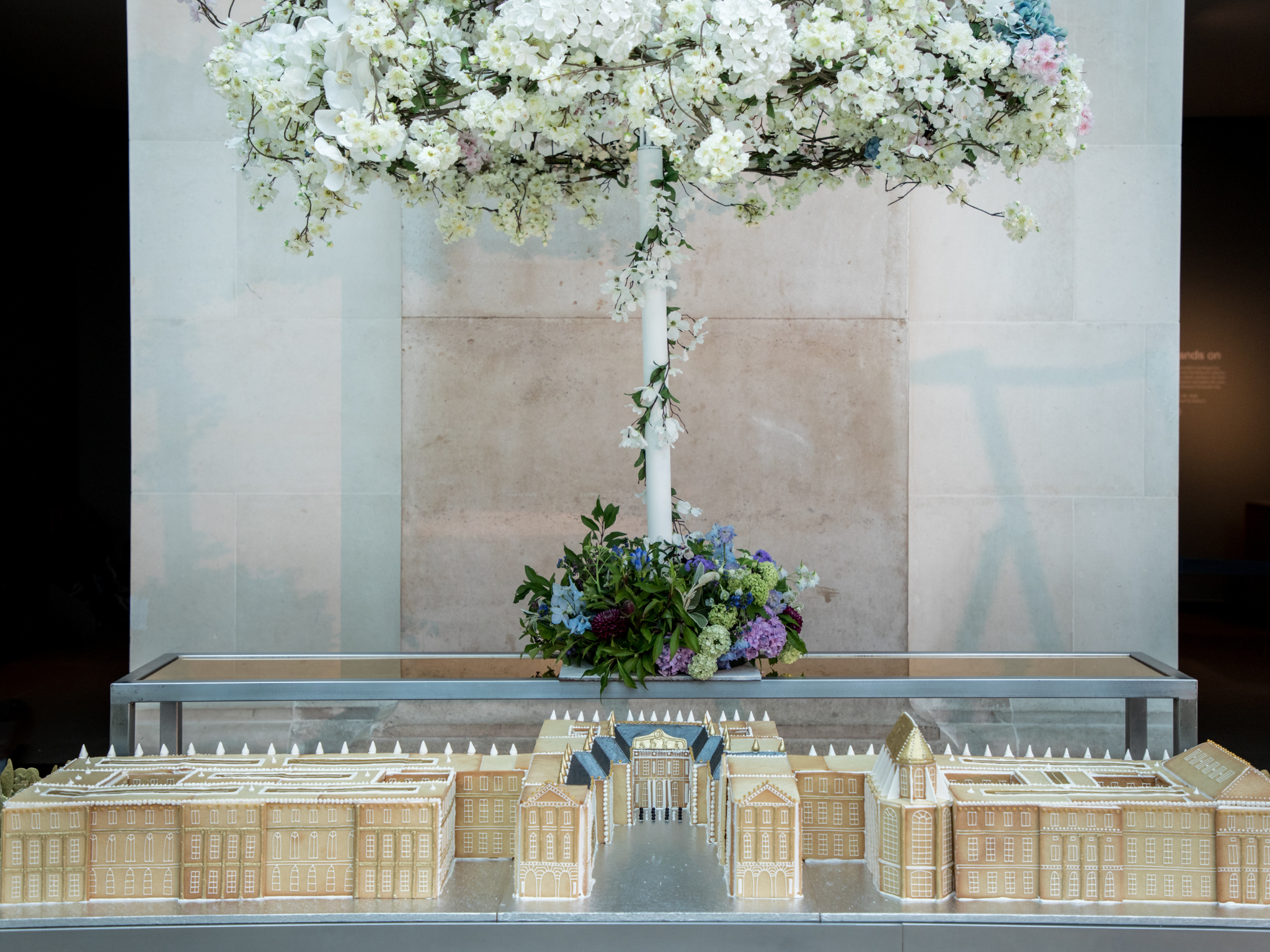 A wide shot featuring a vanilla biscuit replica of the Palace of Versailles, displayed on a large table in the British Museum Great Court, with a decorative floral arrangement above the biscuit building.