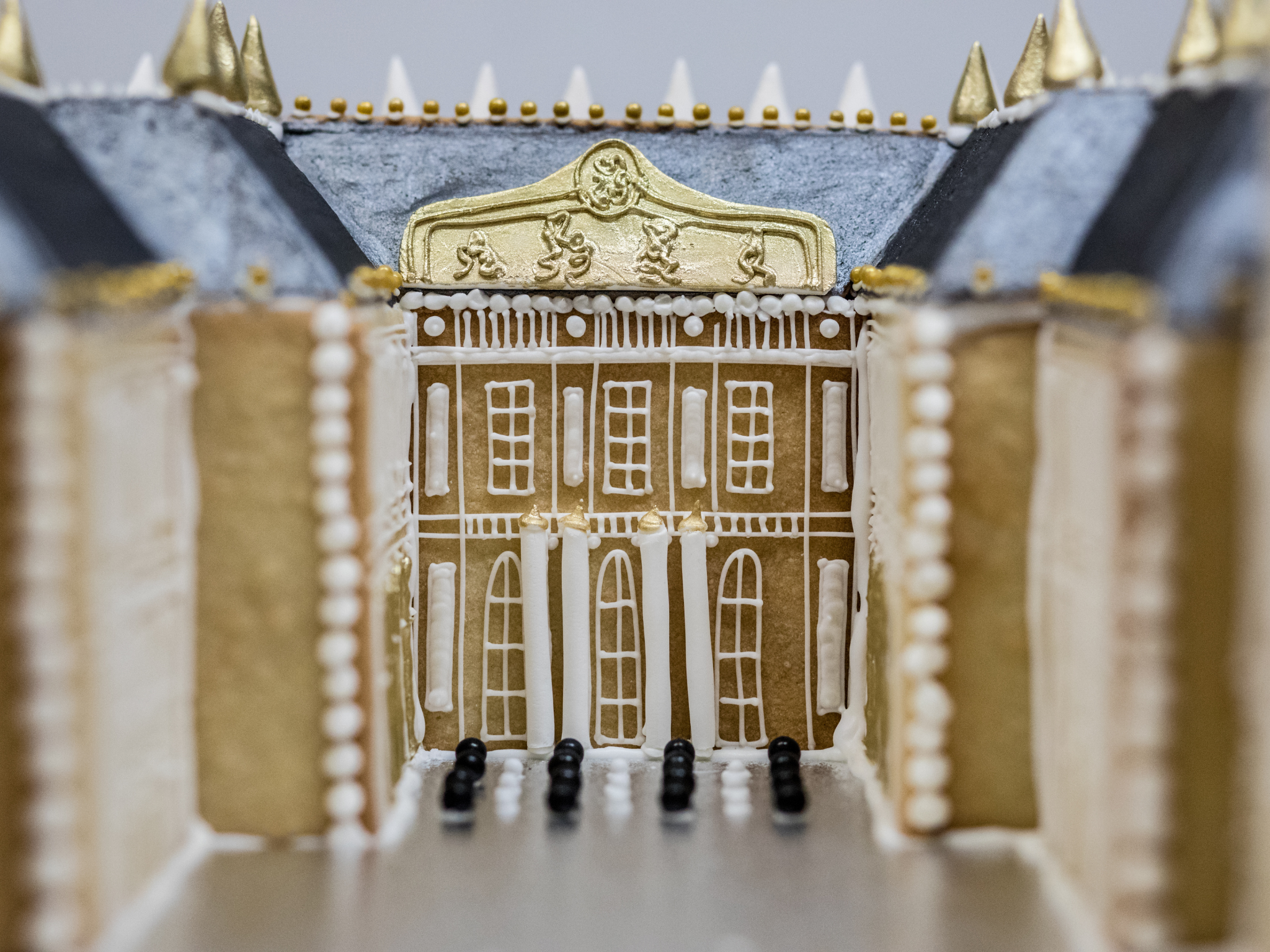 A close-up of a vanilla biscuit replica of the Palace of Versailles, decorated with white and gold icing, featuring detailed windows, columns, and a gold roof with spikes and edible beads.