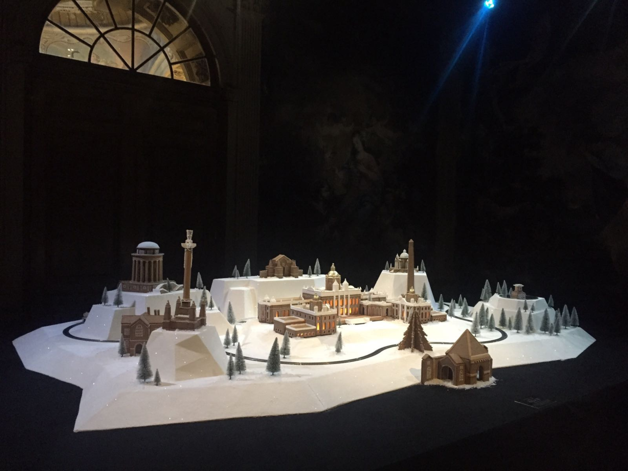 Gingerbread model of Castle Howard with surrounding buildings, monuments, trees and gingerbread train, displayed on a table, spotlit in a dark room.