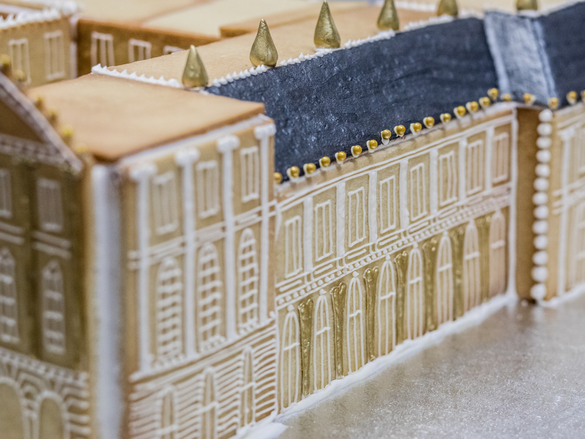 Close-up view of an intricately decorated vanilla biscuit replica of the Palace of Versailles, with white and gold icing patterns along the facade, and gold cone-shaped decorations along the roof.