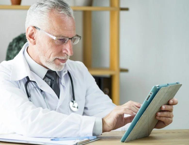 An older male doctor with gray hair, beard, and glasses, wearing a white coat and stethoscope, using a tablet in an office or clinical setting.