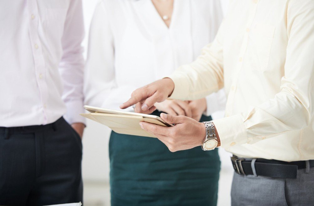 Three people in professional attire discussing documents or a tablet in a business setting.