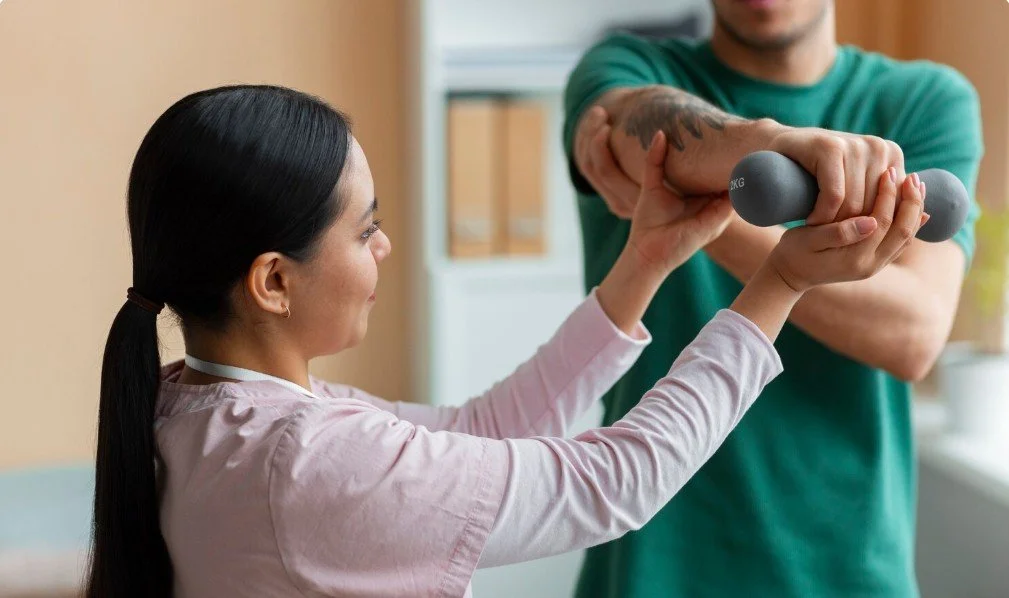 A woman in a pink shirt is holding a grey dumbbell with assistance from a man in a green shirt, indoors.