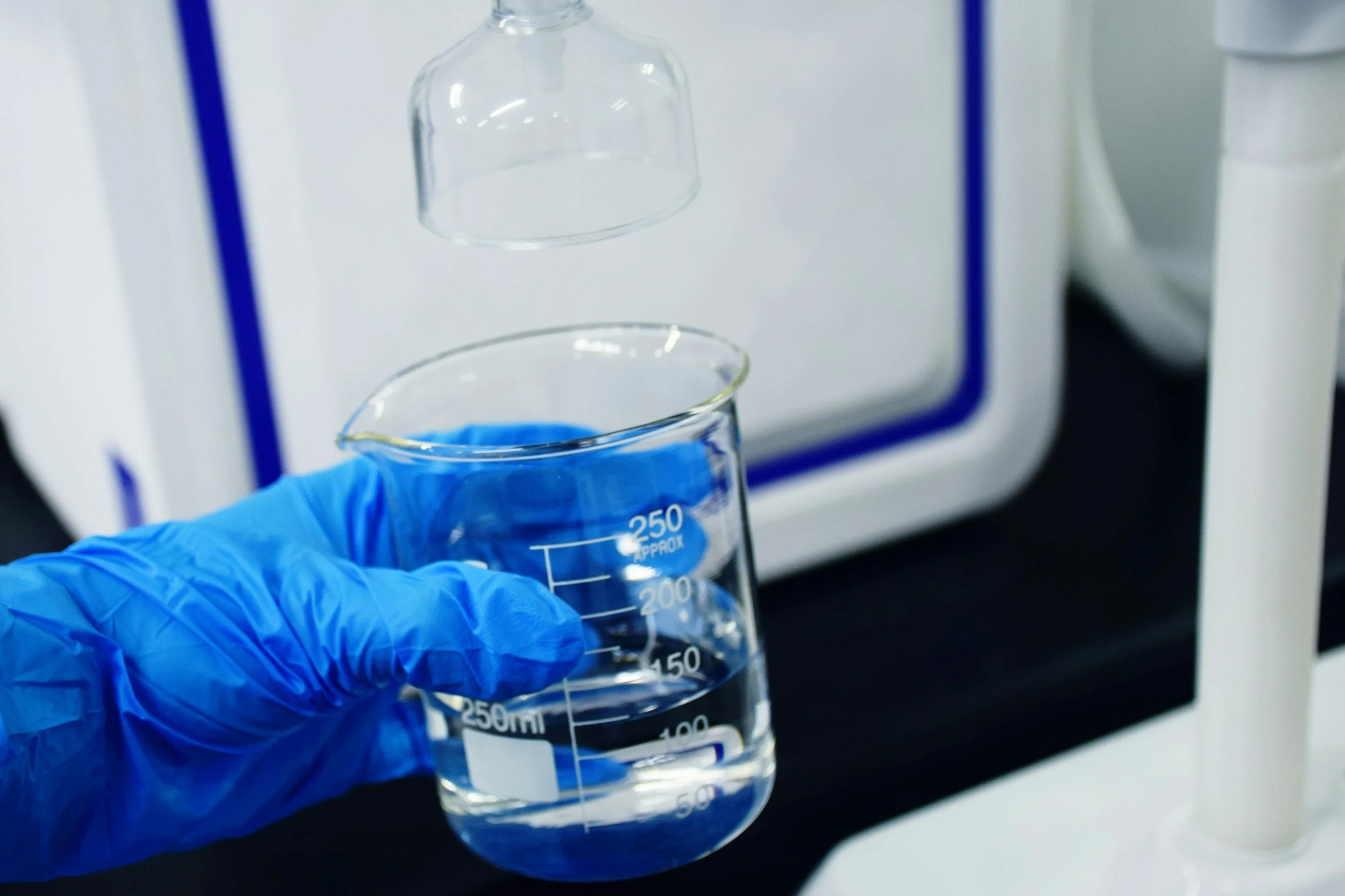 Person wearing blue gloves holding a clear measuring beaker with blue liquid in a laboratory setting.