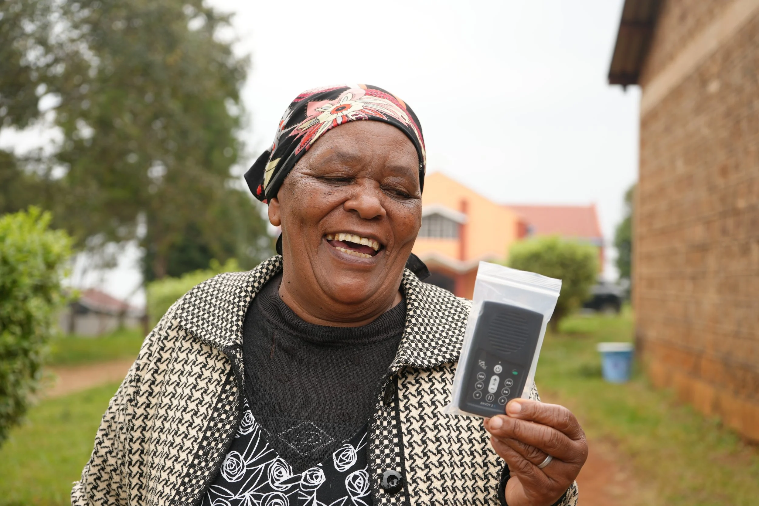 An elderly woman smiling and looking at an audio Bible she is holding in her hand, outside with trees and buildings in the background.