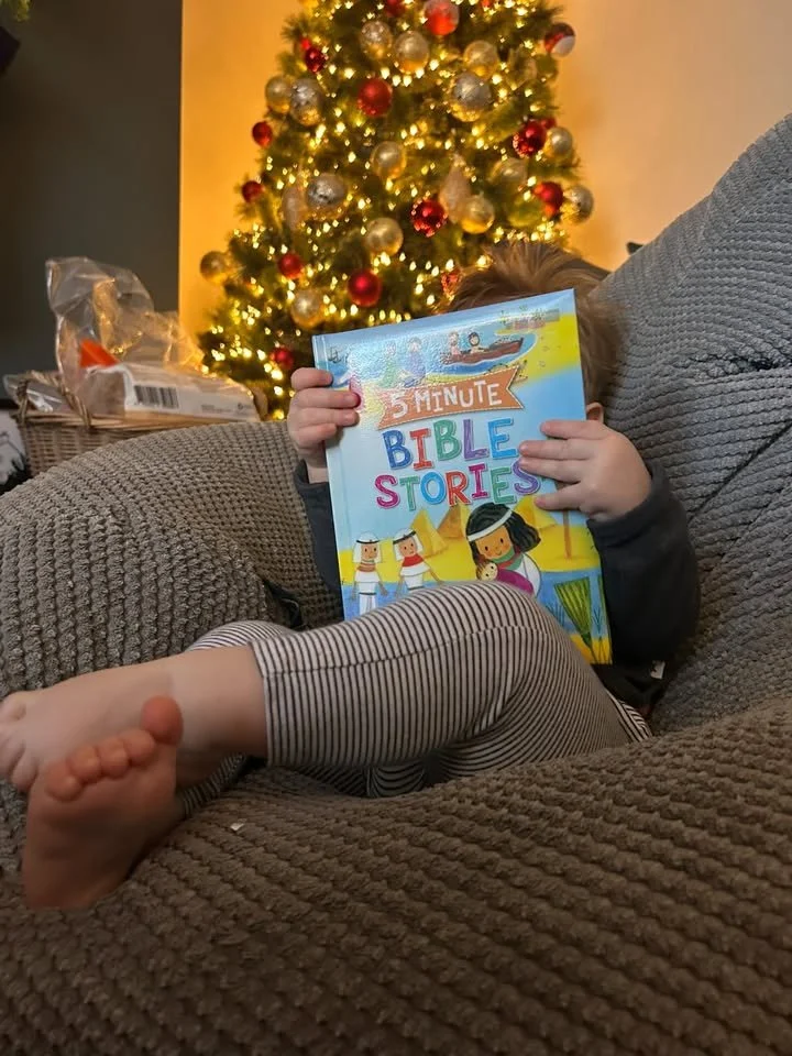 A young child sitting on a brown textured couch holding a colorful children's book titled '5 Minute Bible Stories' in front of a decorated Christmas tree with red, gold, and silver ornaments and lights.