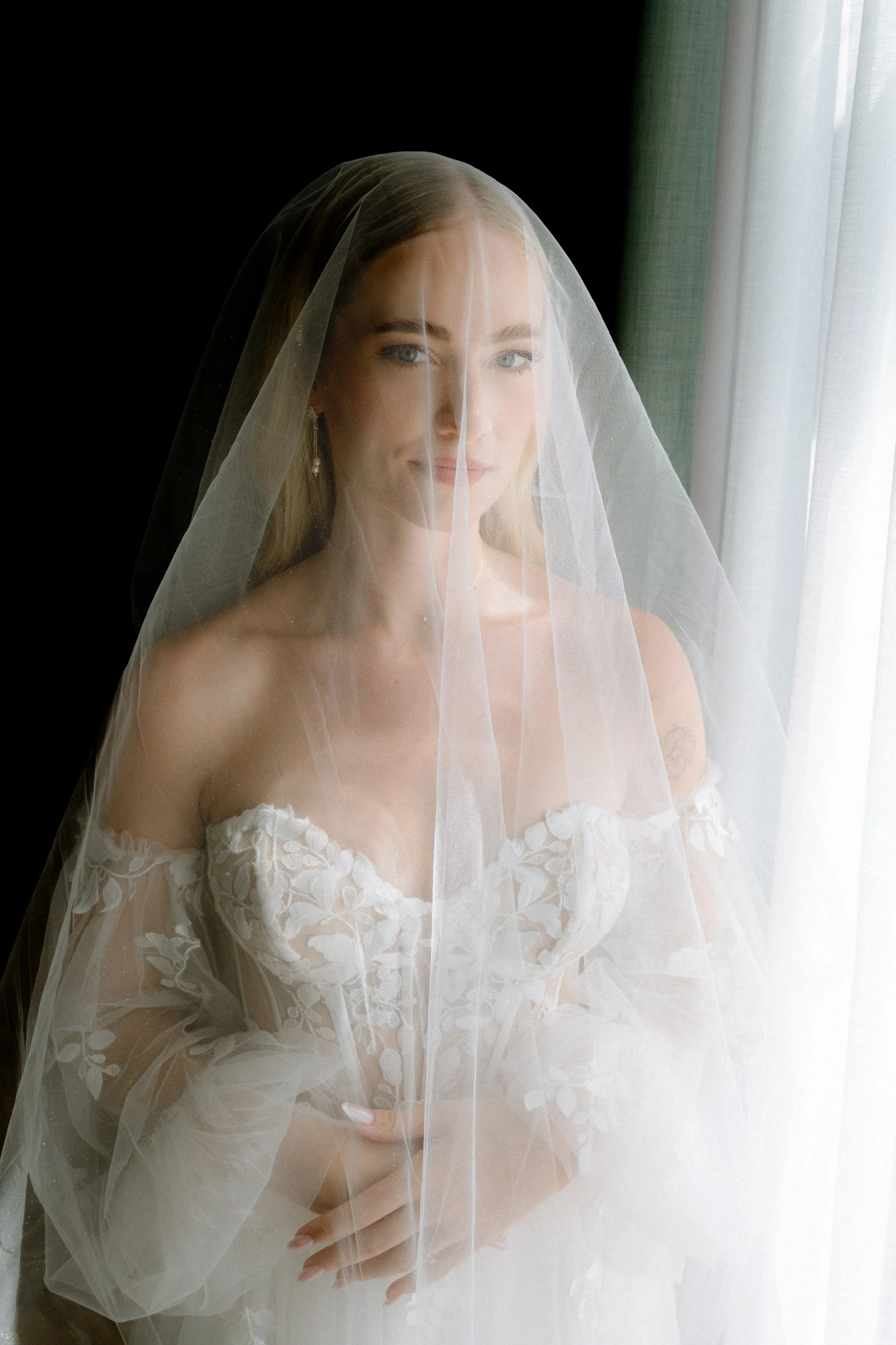 A woman in a wedding dress and veil standing by a window with sheer curtains, looking at the camera.