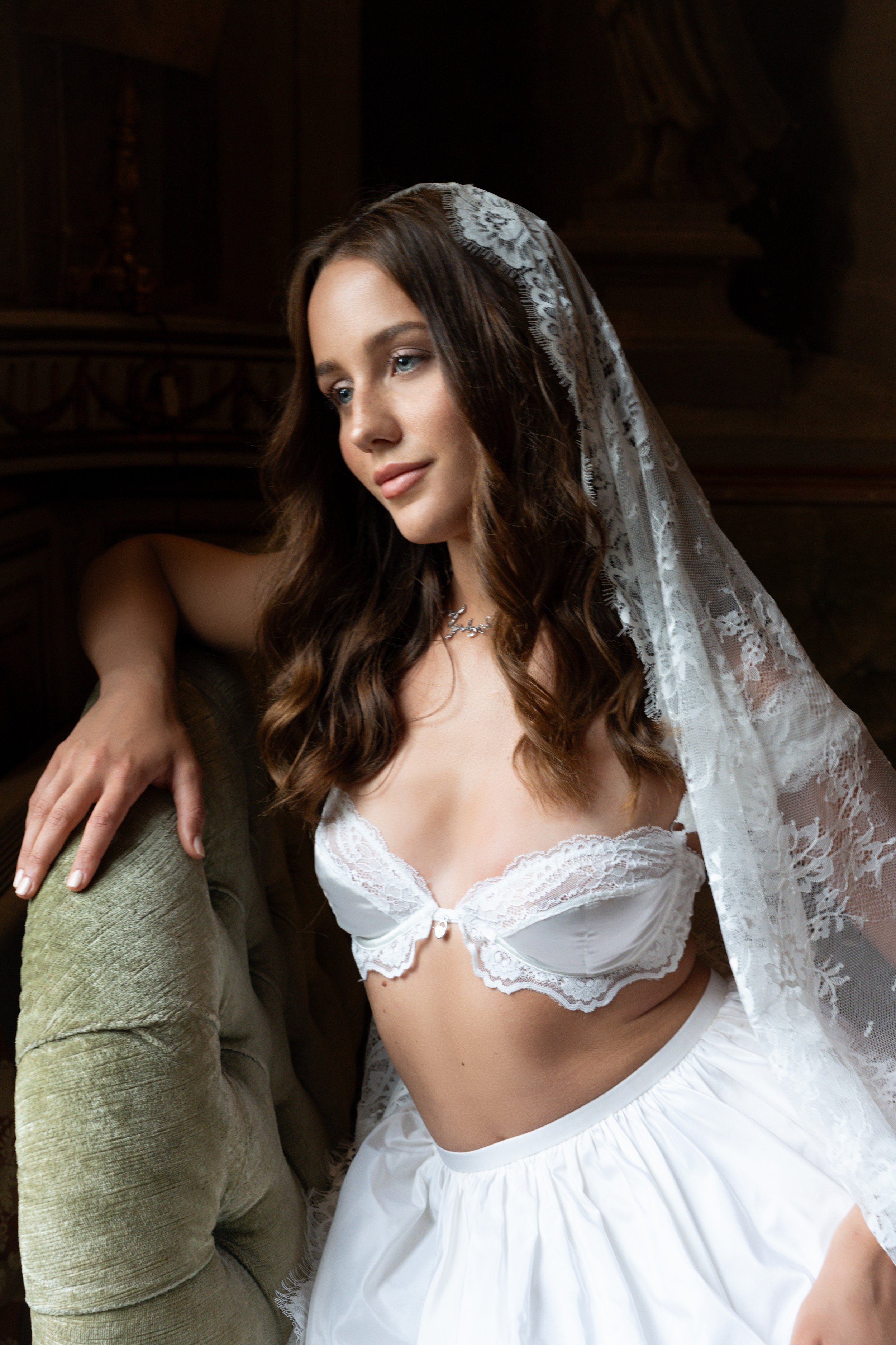 A young woman in white lace lingerie and a satin skirt, wearing a lace veil, seated on an armchair with a wooden background.