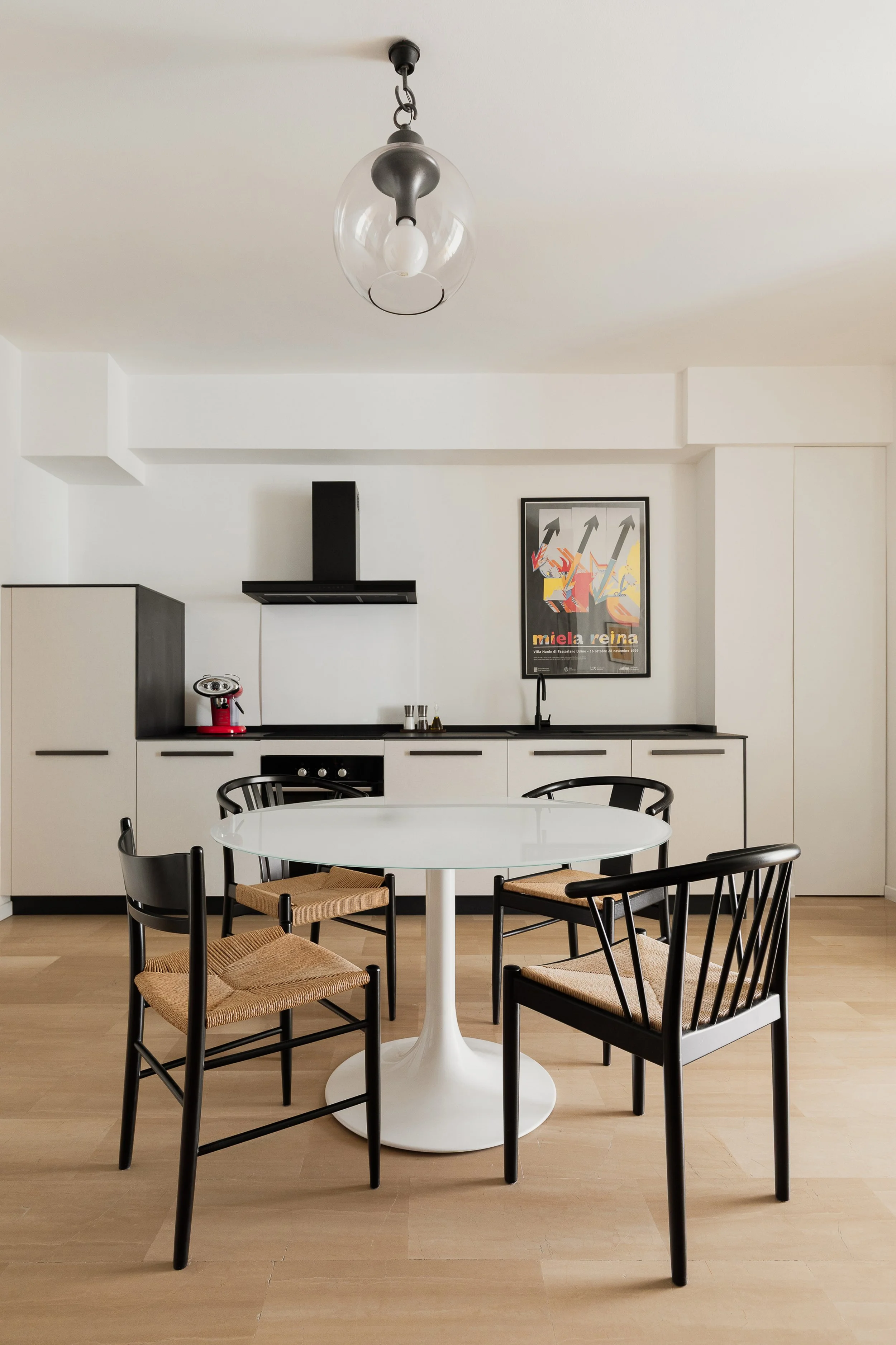Cucina moderna con tavolo tondo bianco e quattro sedie nere con sedili in paglia, armadi bianchi e piano di lavoro nero, lampadario sospeso e poster con frecce e testo colorato.