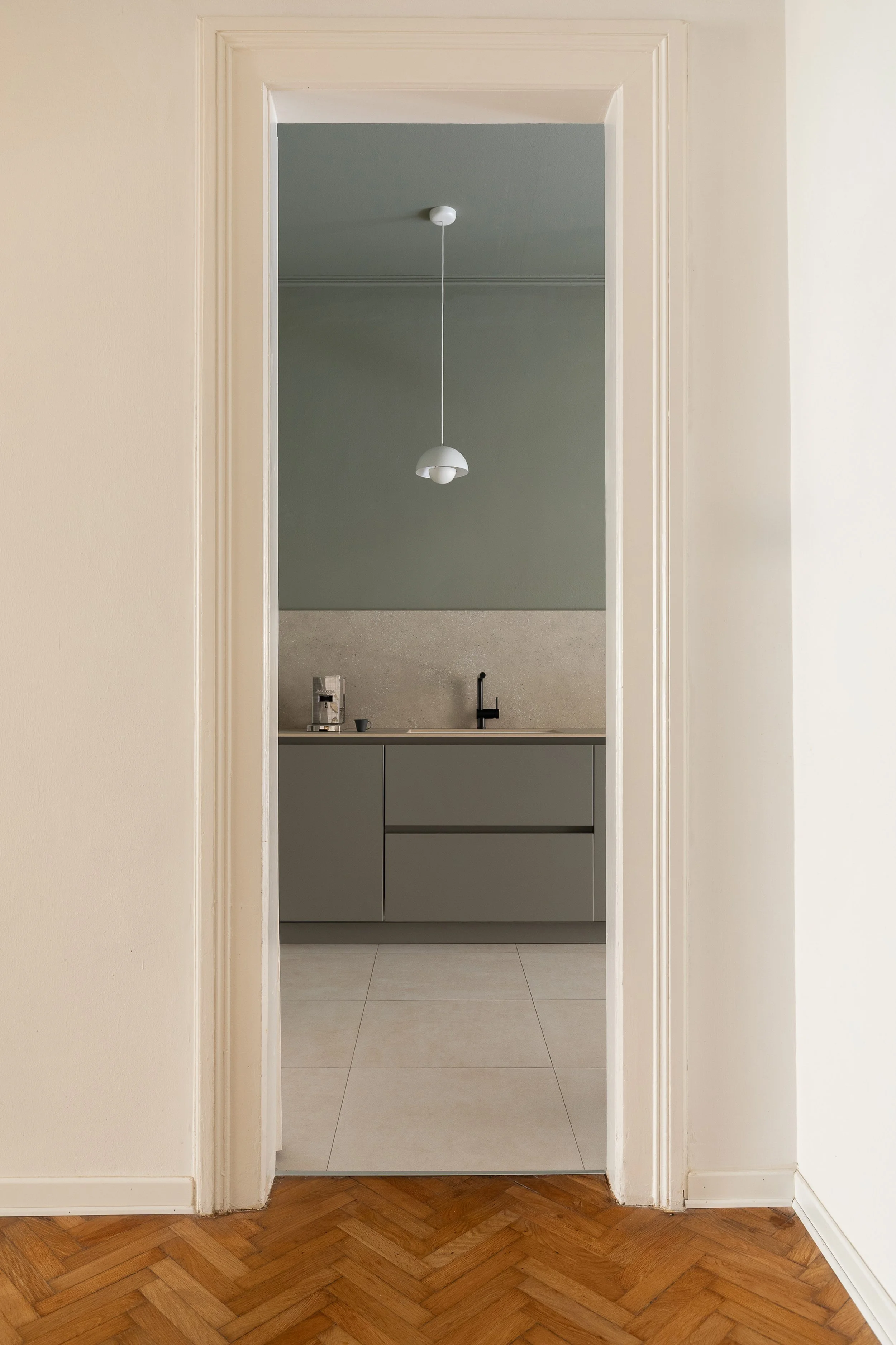 Vista di una cucina moderna con mobili grigi e pensile, lavello nero, erogatore di acqua e lampadario bianco sospeso, visibile attraverso una porta bianca.