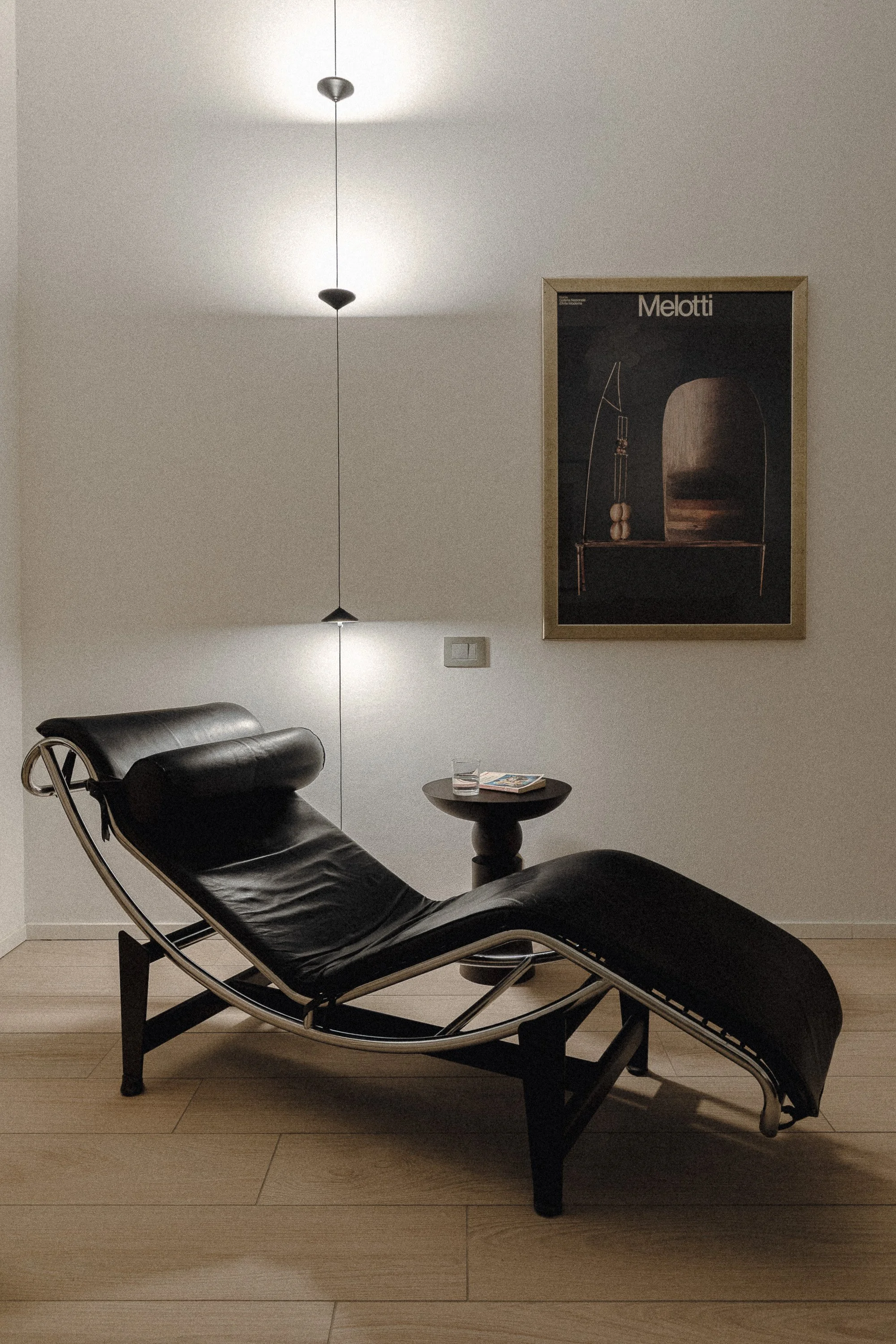 Sedia a chaise longue nera in stile modernista in una stanza con pareti bianche, un quadro astratto con la scritta 'Melotti', un'illuminazione a sospensione e un tavolino con una rivista e un bicchiere.
