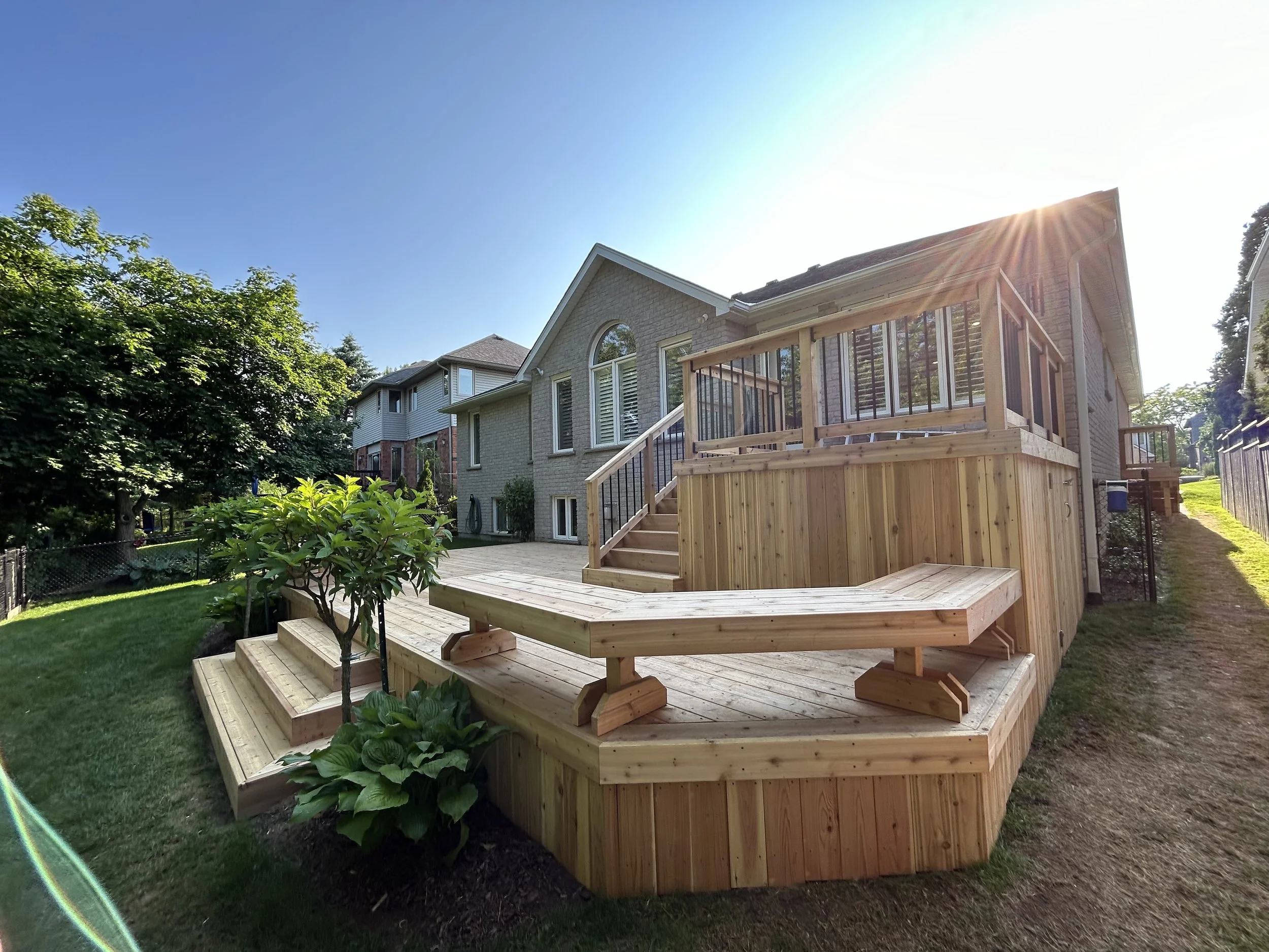 Backyard view showing a newly built wooden deck with stairs and a raised platform attached to a house, with a garden and neighboring houses in the background, under a clear blue sky with sunlight.