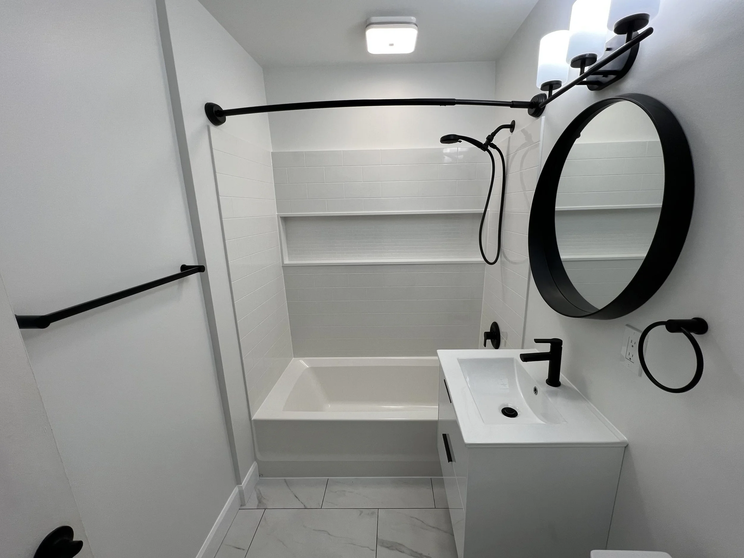 Modern white bathroom with black fixtures, featuring a round mirror, wall-mounted lights, white vanity with sink, black faucet, bathtub with showerhead, white tiled walls, and marble floor.