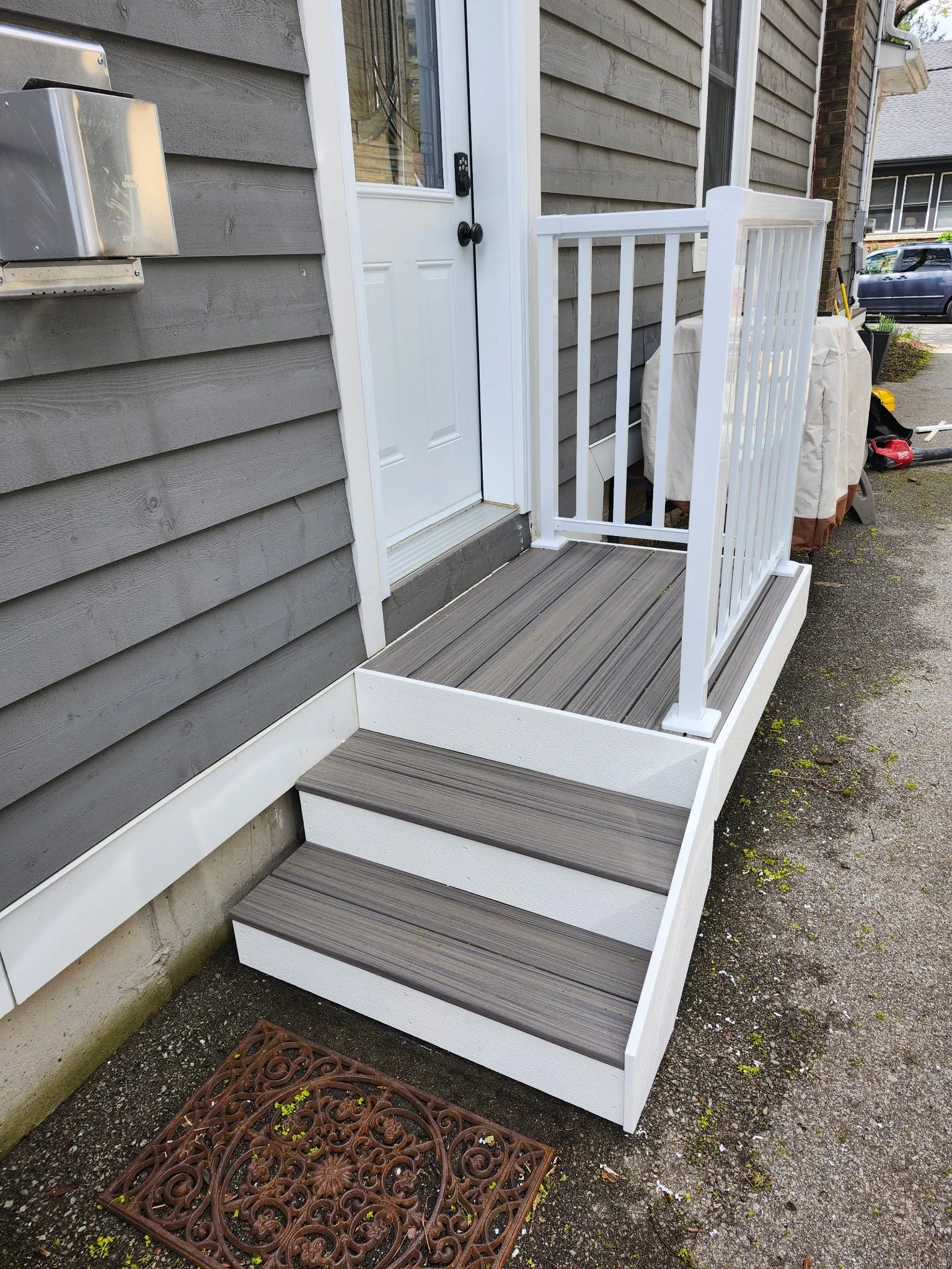 A small porch with three steps leading up to a door, featuring gray wood flooring and white railings, next to a gray house with horizontal siding.