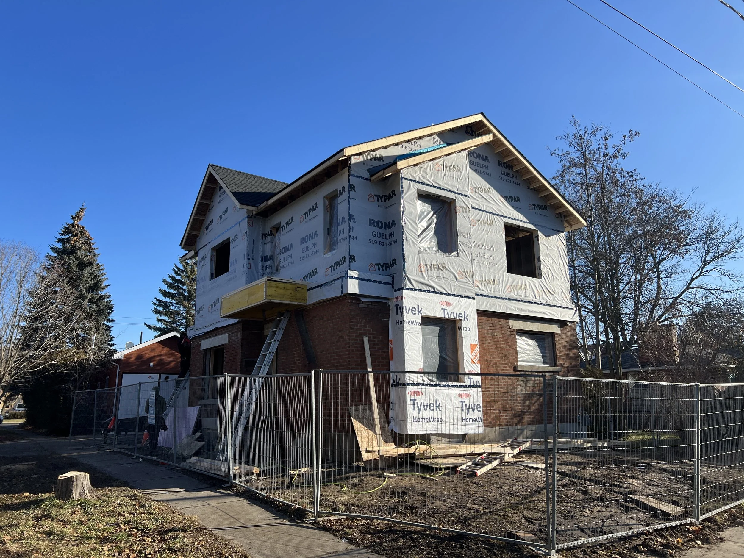 Two-story house under renovation with Tyvek insulation wrap, a ladder, and scaffolding, surrounded by a metal fence.