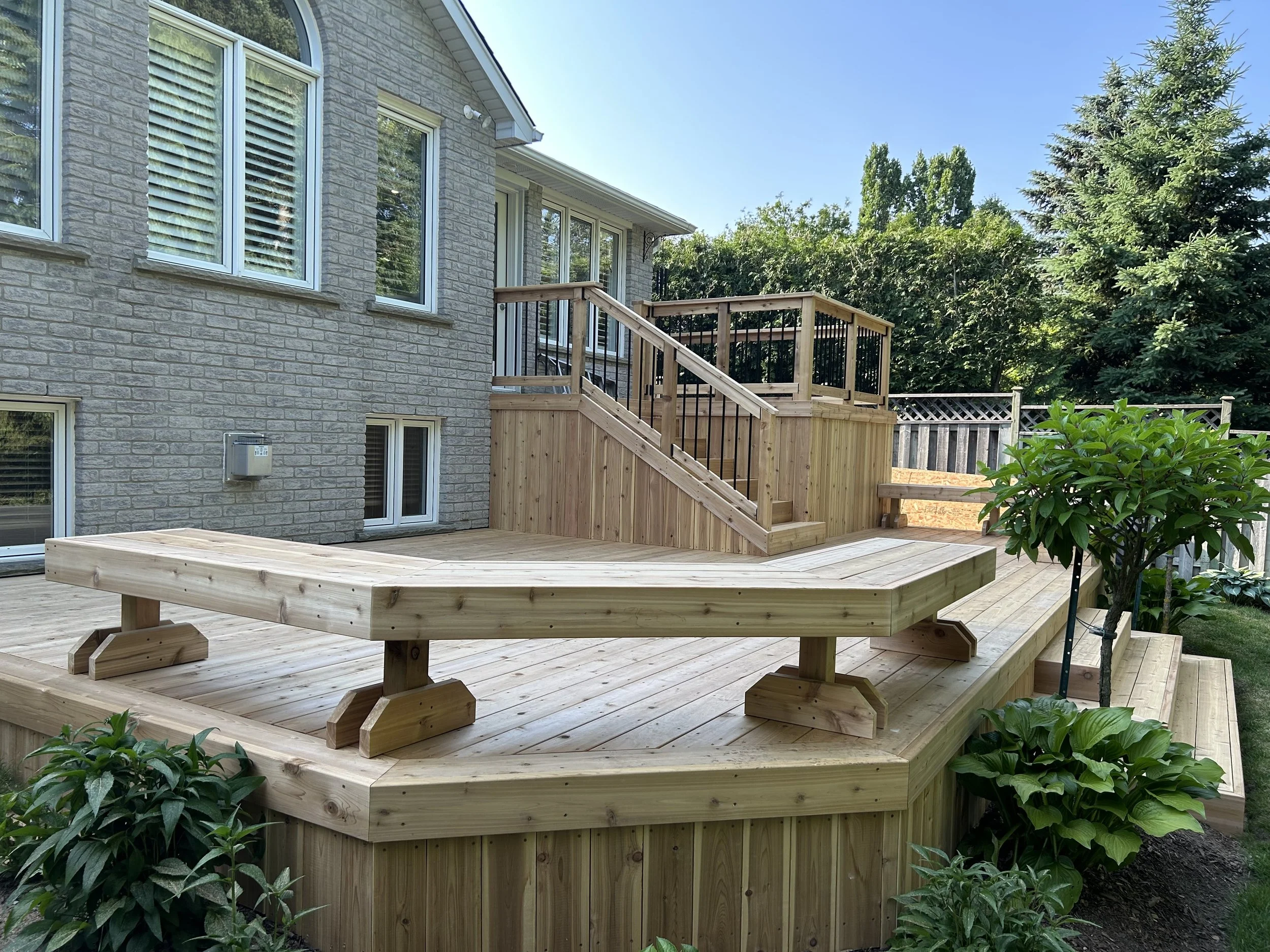 New wooden deck with stairs and built-in bench around the backyard of a house with brick exterior, surrounded by green trees and plants.