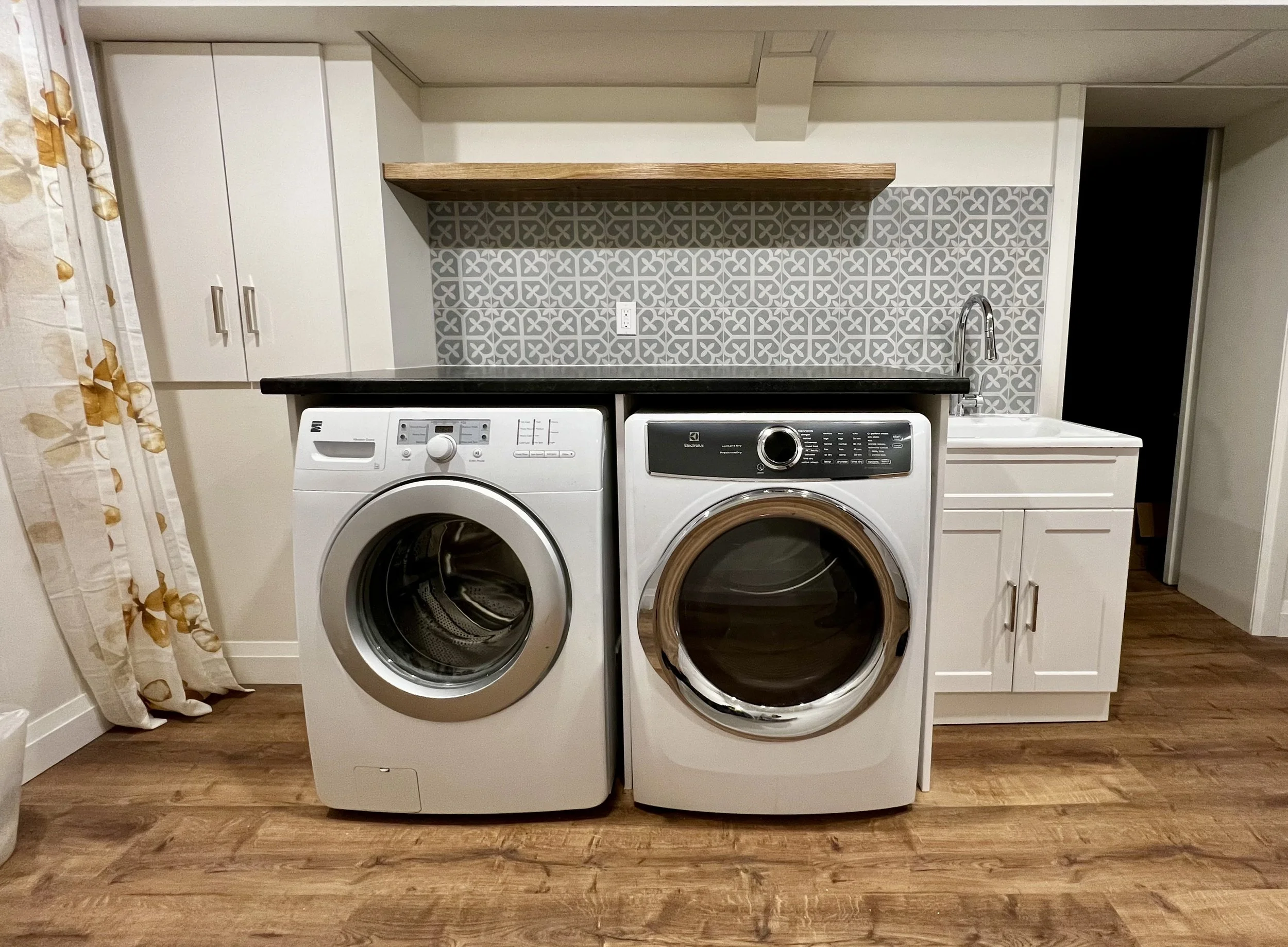 Washer and dryer in a laundry room with white cabinets, a wooden shelf, patterned gray tile backsplash, and a sink with a faucet.